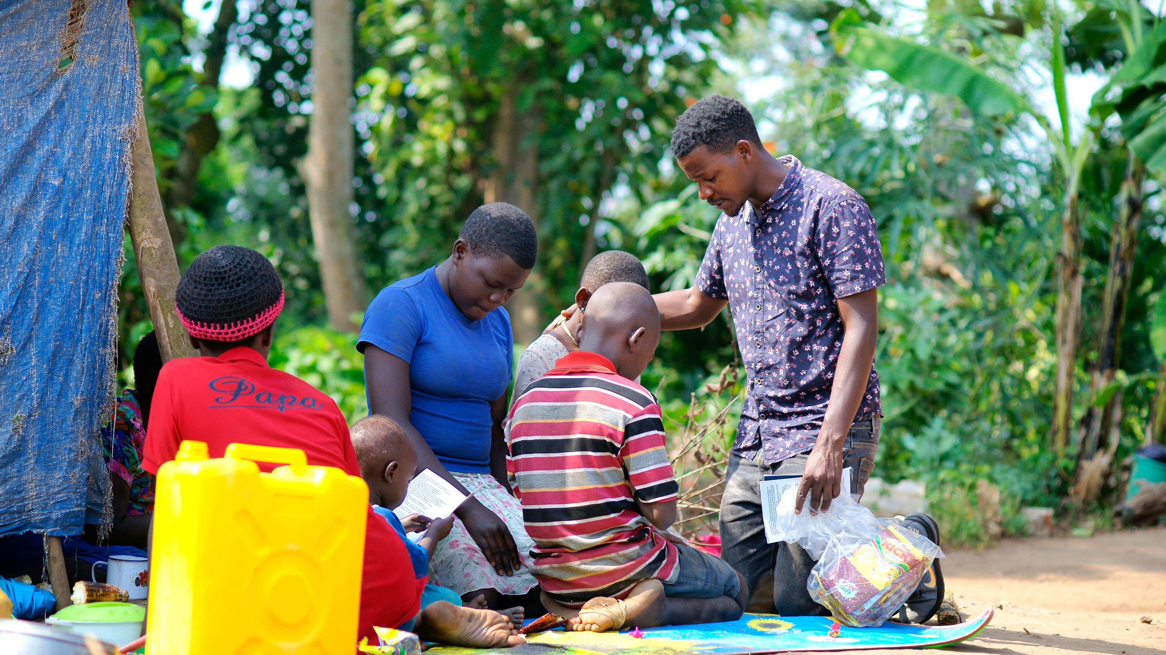A group gathers outdoors near a blue tarp shelter as a man distributes food, a yellow jerry can nearby.