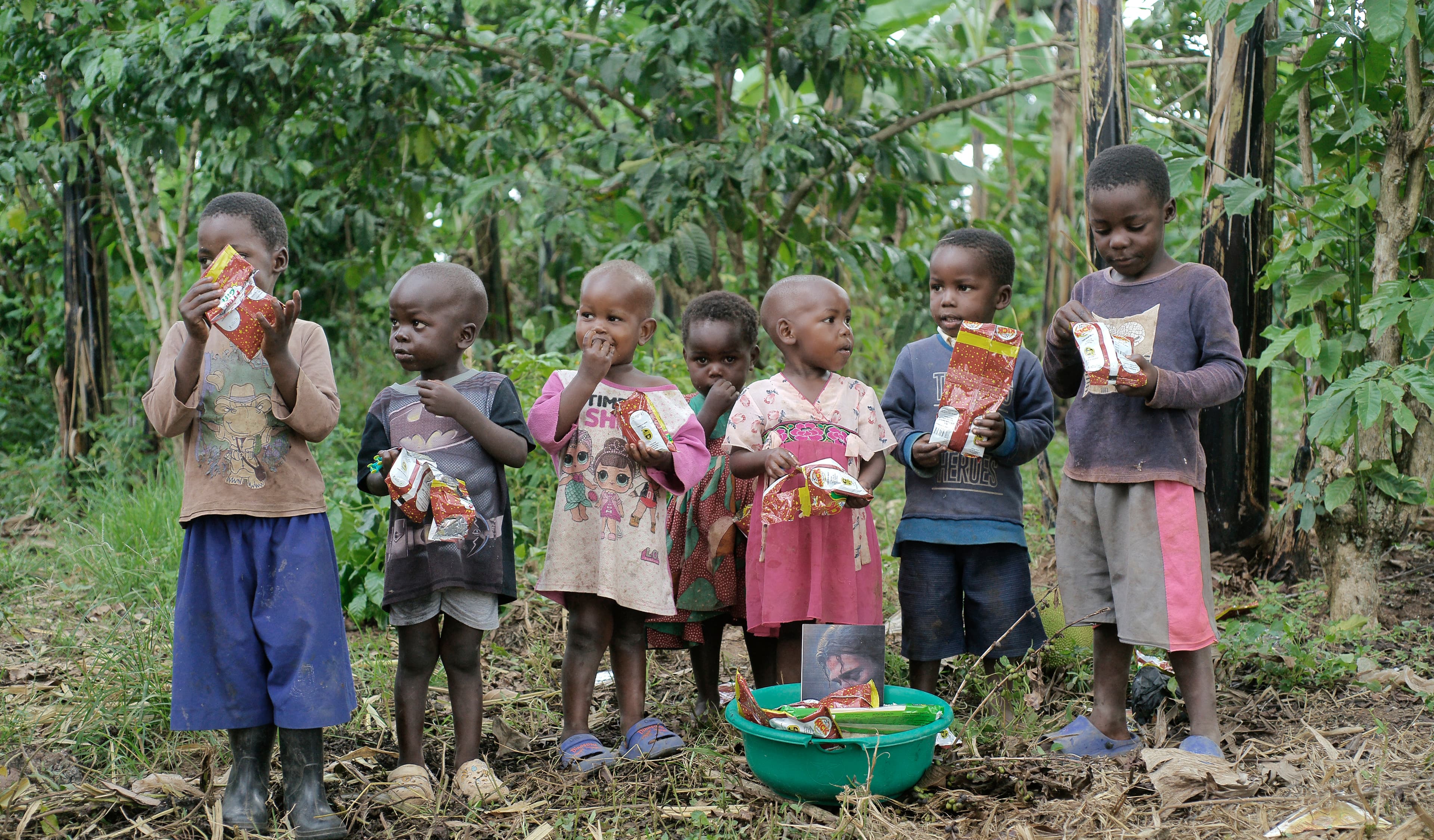 A group of children stand together outdoors eating snacks from packets after a food distribution.