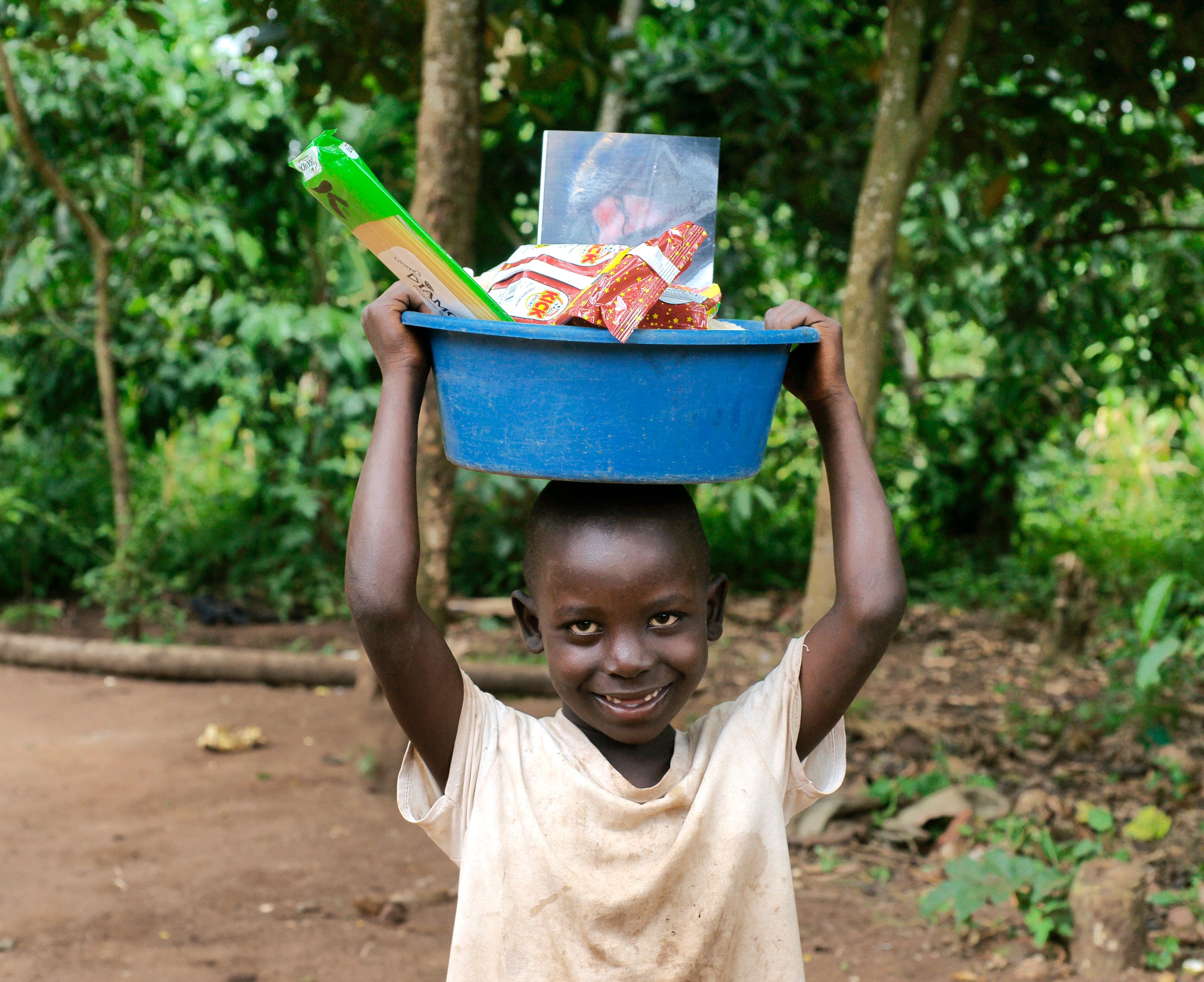 A girl carries a blue basin of food items on her head while walking along a dirt path.