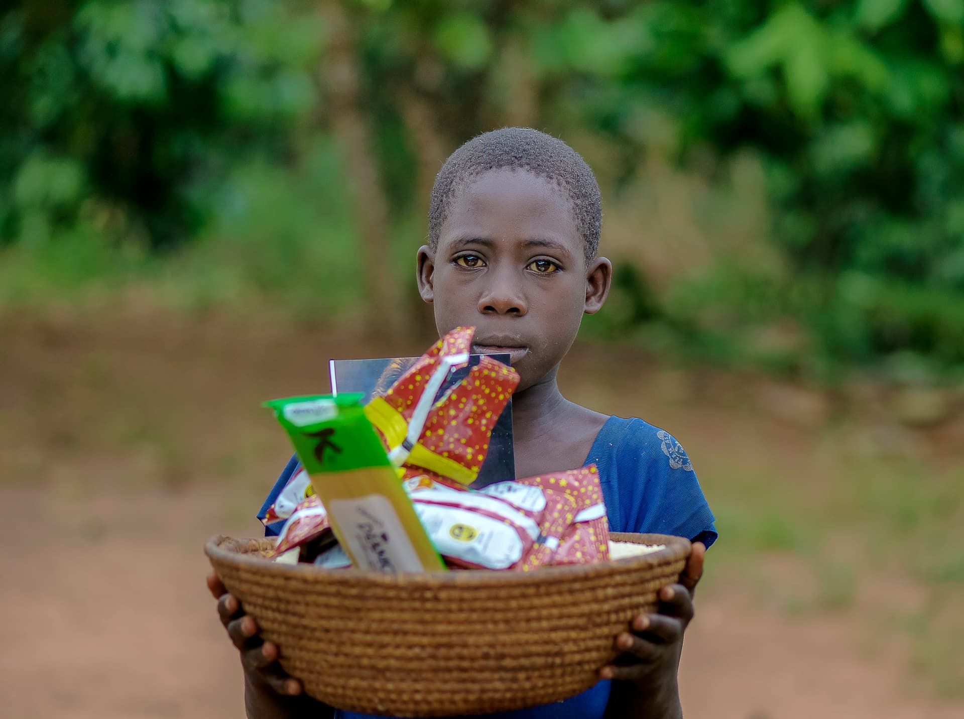 A boy holds a woven basket filled with snack packets, looking at the camera with a worried expression.