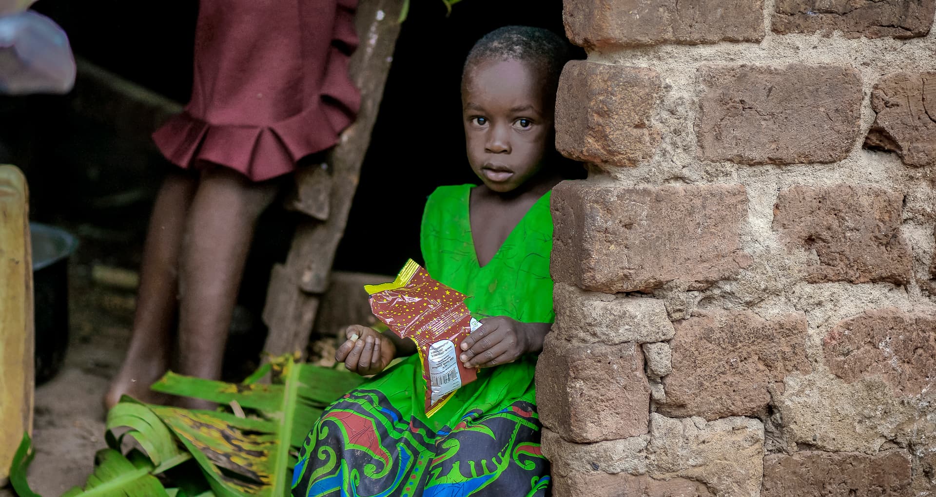 Two children in colorful clothing stand by a brick wall, one holding packaged food items.
