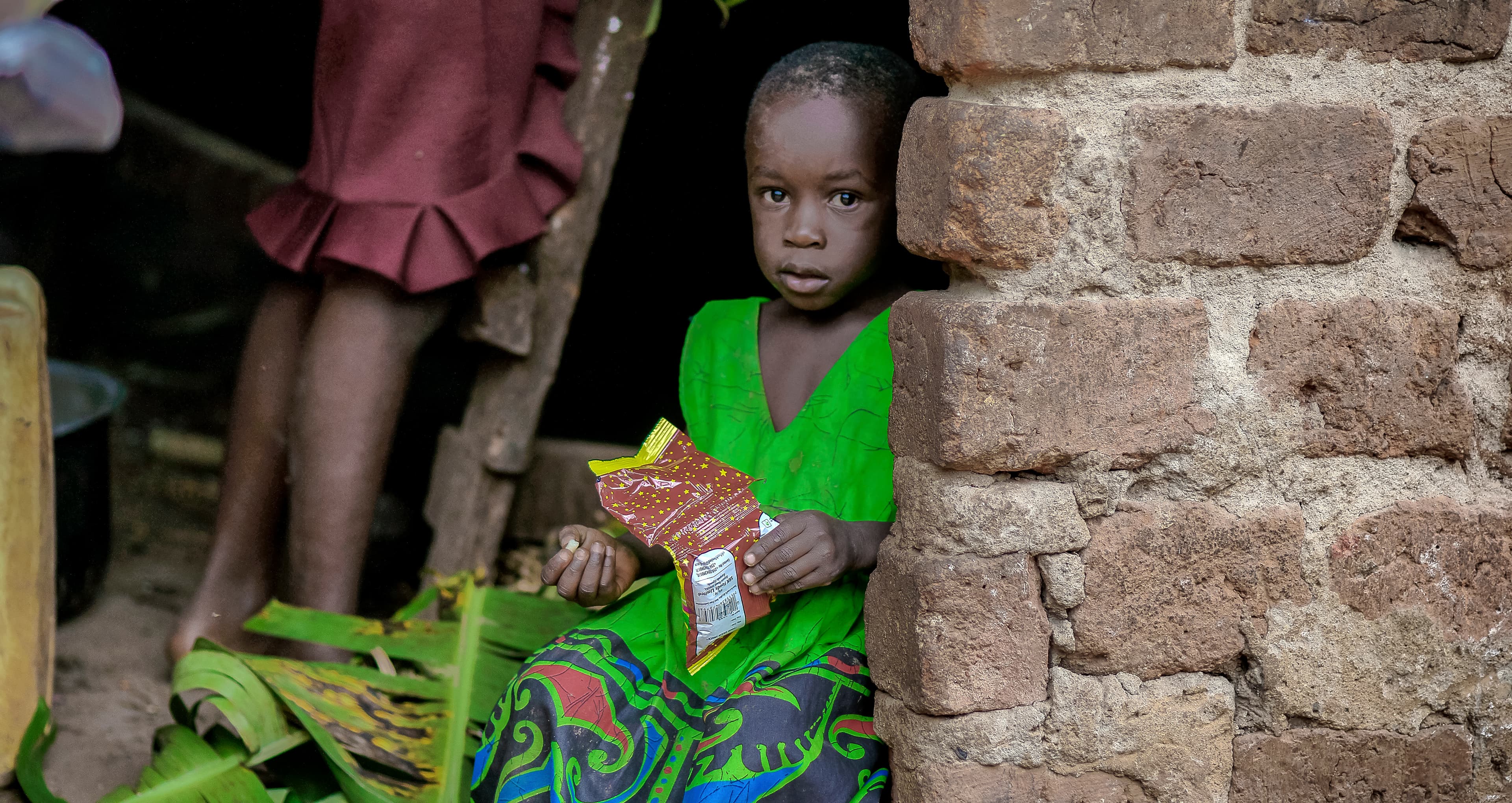 Two children in colorful clothing stand by a brick wall, one holding packaged food items.