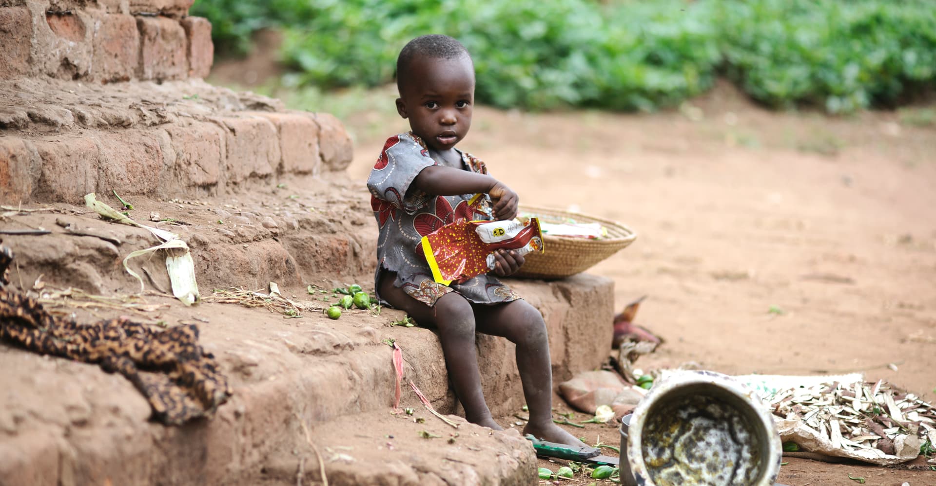 A young child sits on the ground near a brick wall holding food items beside a metal plate.