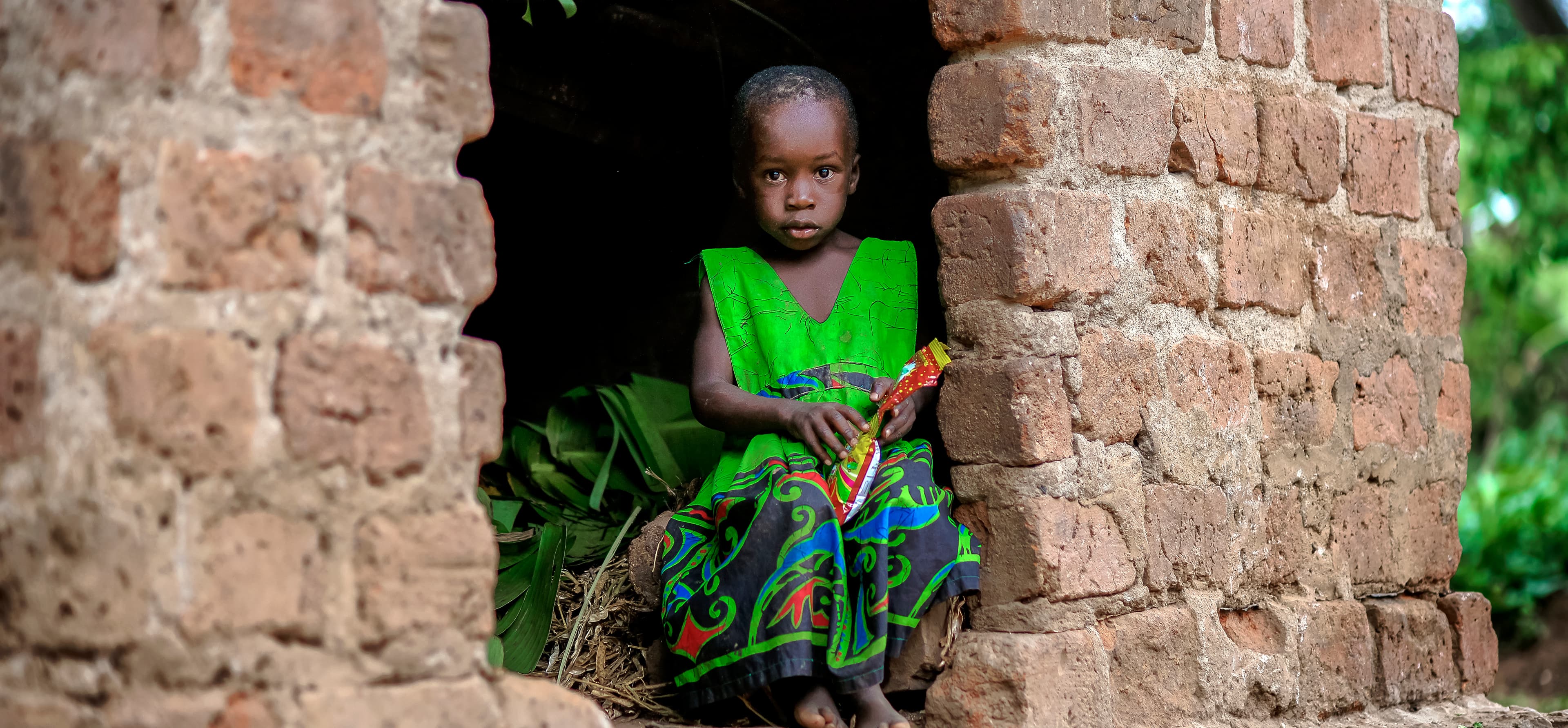 A child in a green outfit stands beside a brick wall or structure.