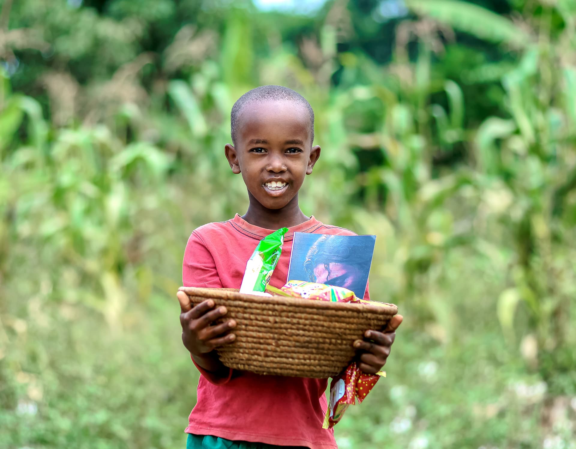 A boy smiles while holding a woven basket filled with grocery and snack packets outdoors.