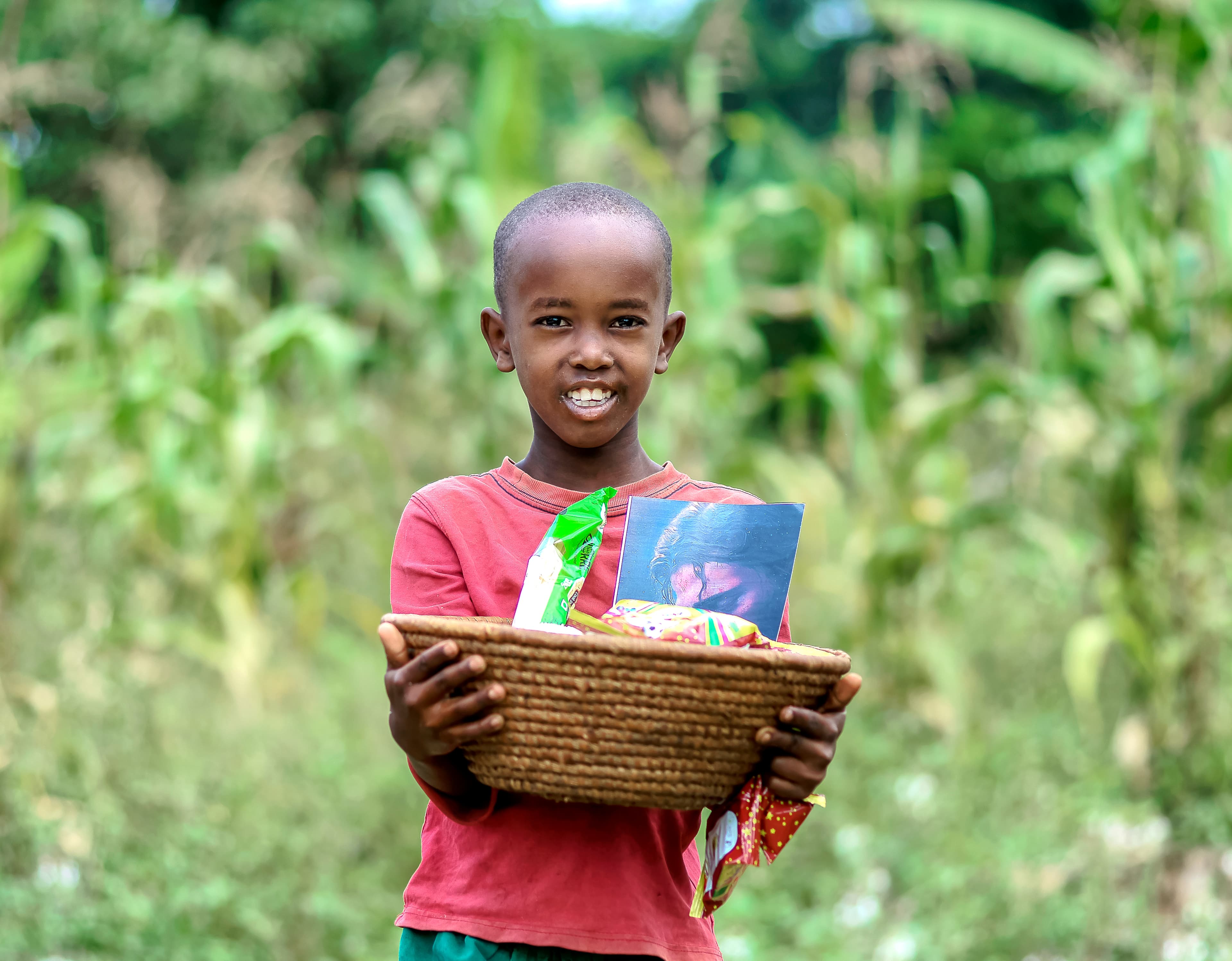 A boy smiles while holding a woven basket filled with grocery and snack packets outdoors.