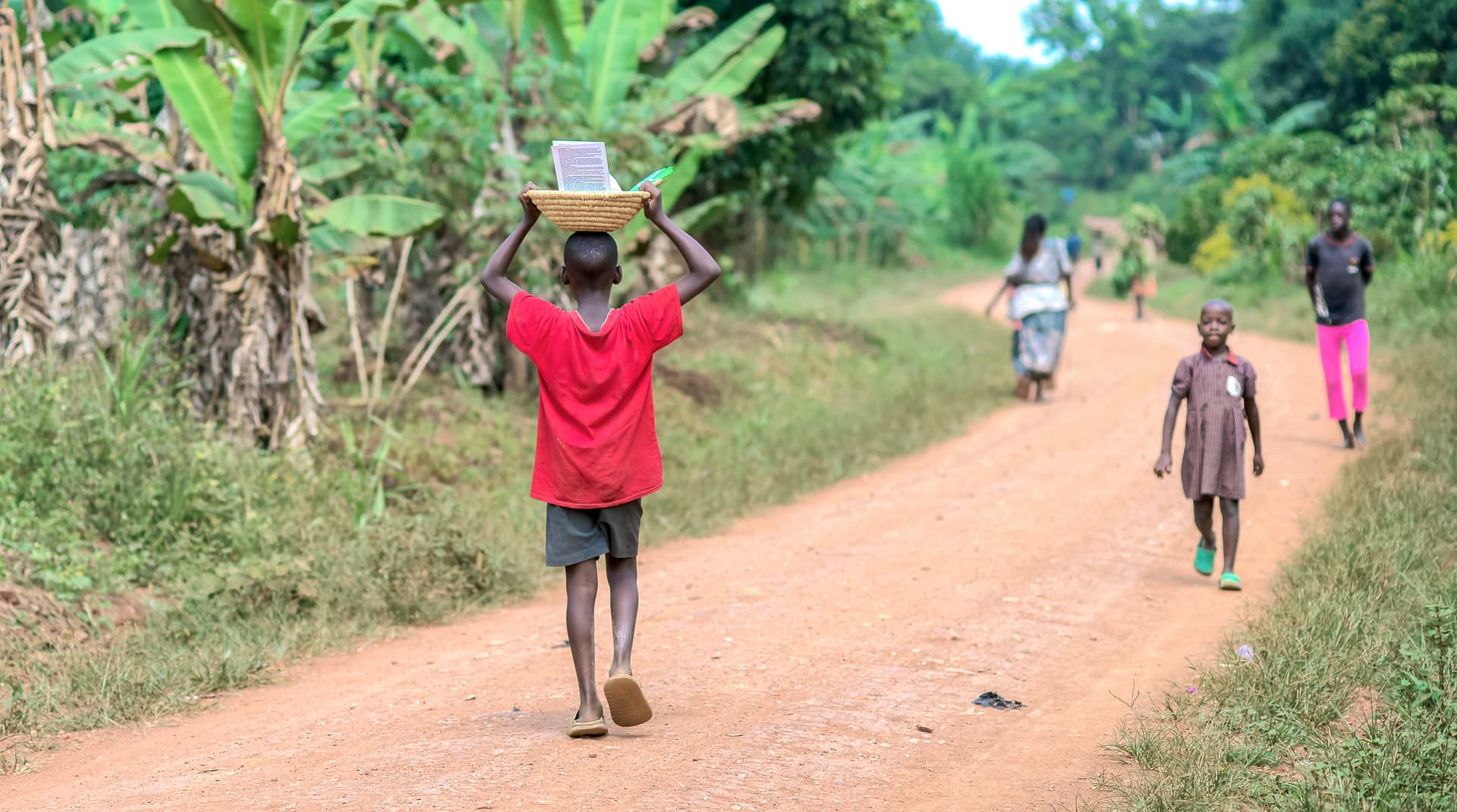 A boy in a red shirt walks down a dirt road carrying items, other people visible behind him.
