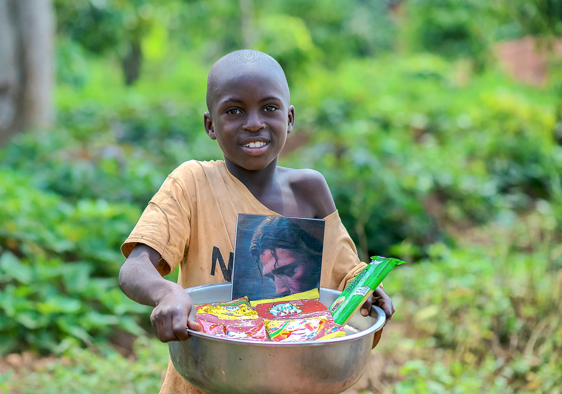 A boy smiles while holding a metal bowl filled with grocery items.