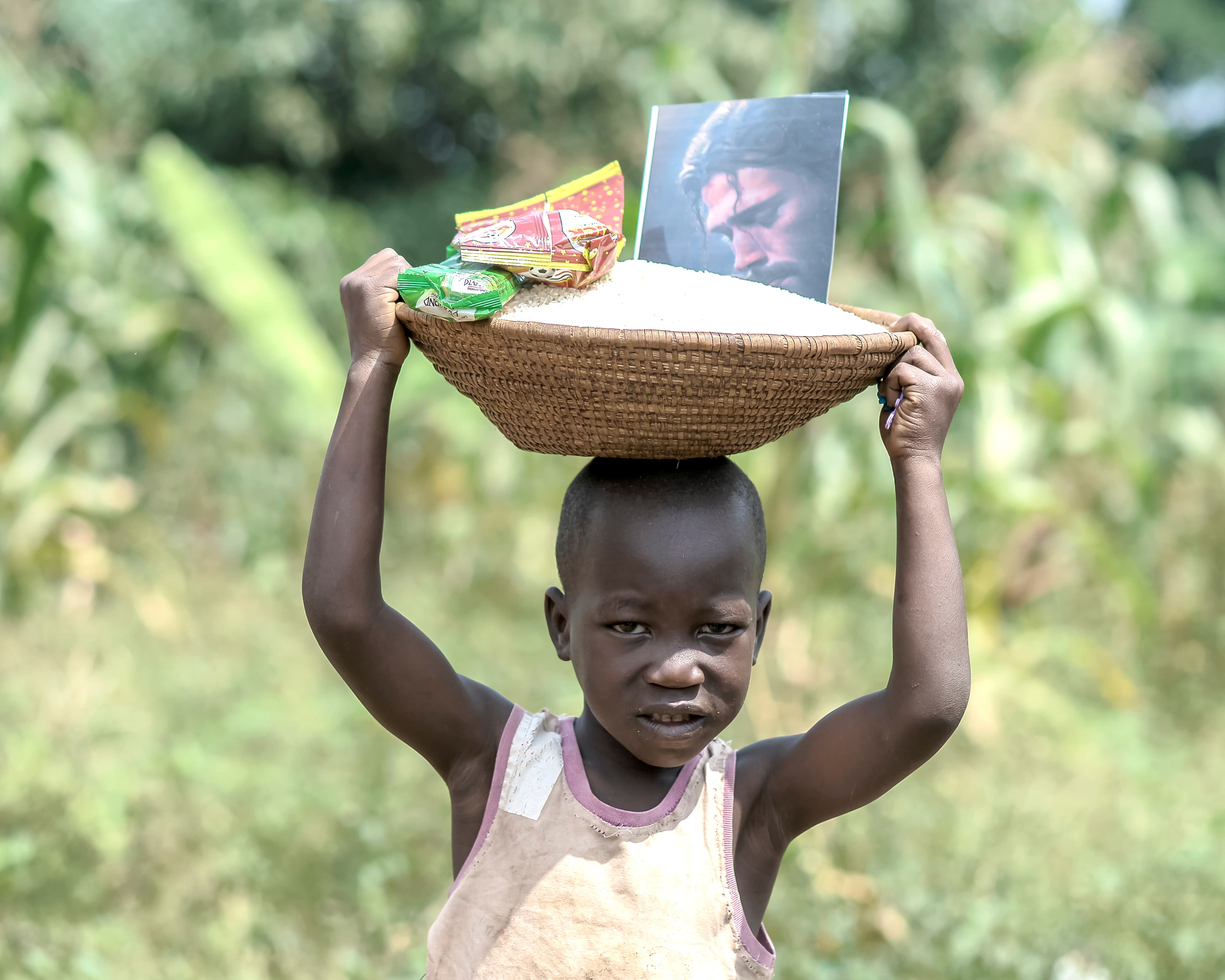 A boy carries a woven basket of snack packets on his head while another child stands nearby.