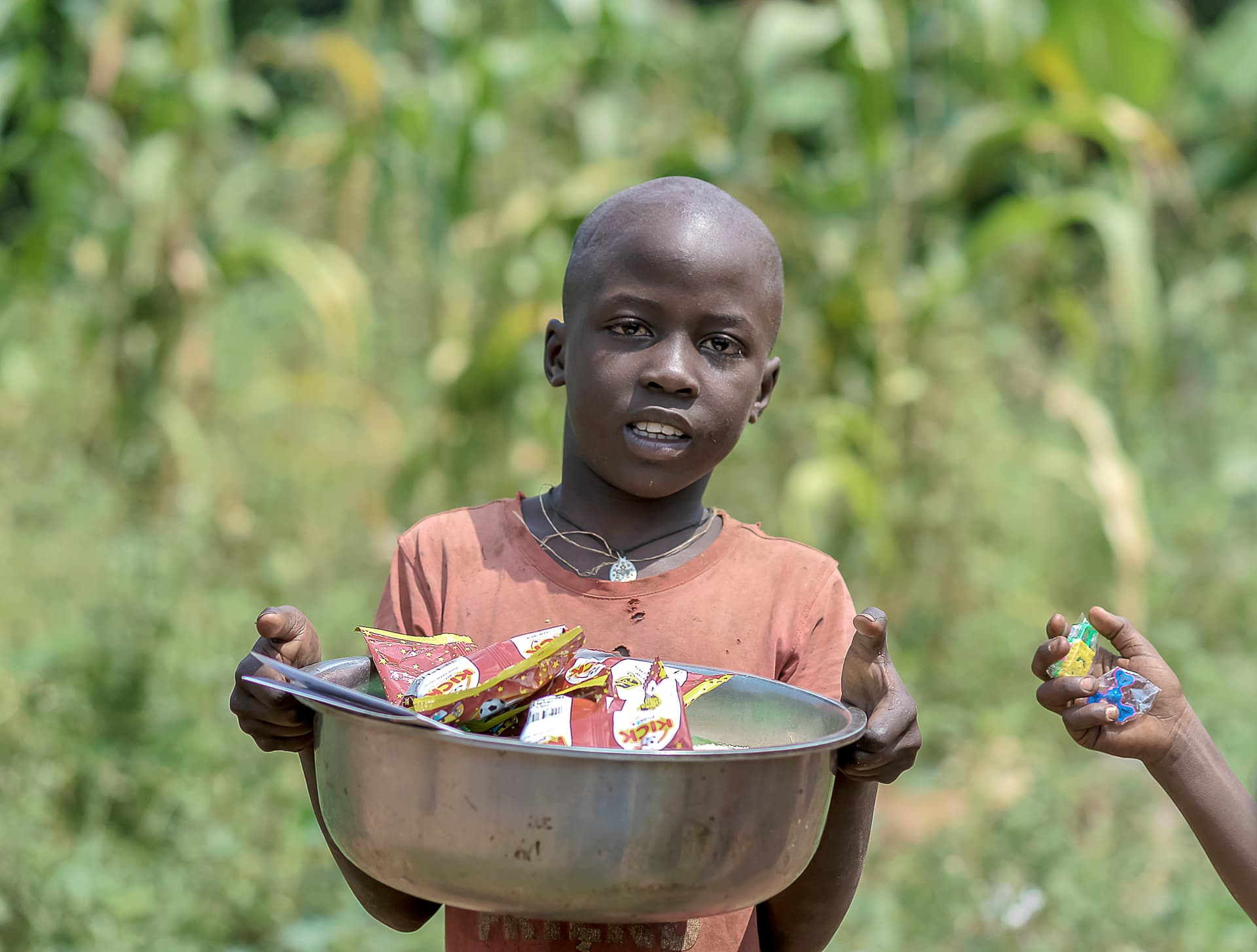 A boy in an orange shirt holds a metal bowl of grocery items and looks at the camera.