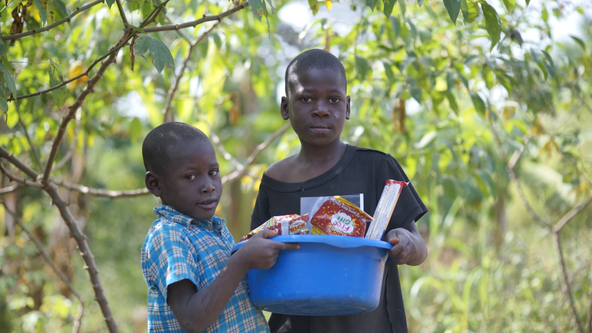 Two boys standing together outdoors, one holding a blue basin of food and papers