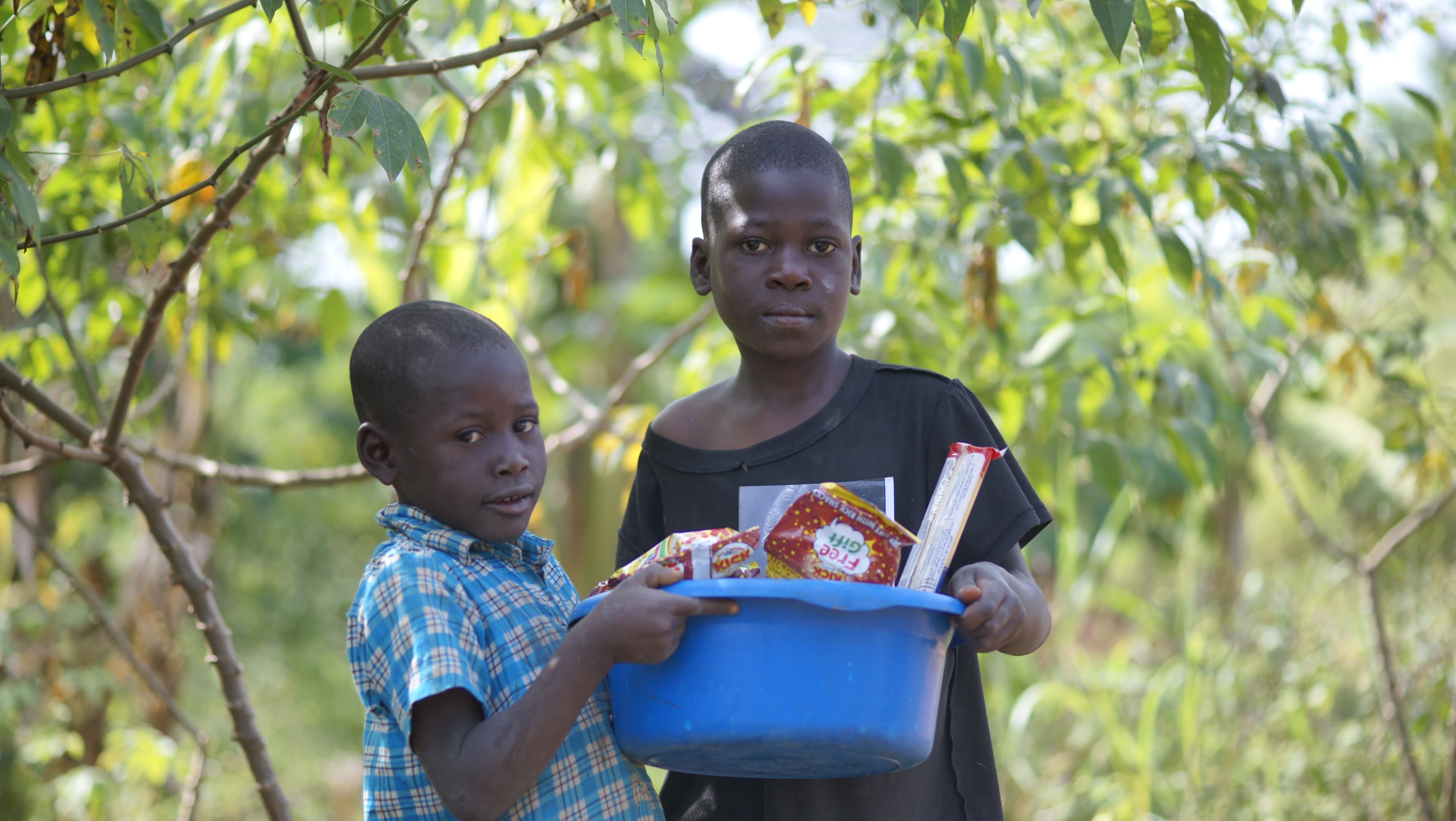 Two boys standing together outdoors, one holding a blue basin of food and papers