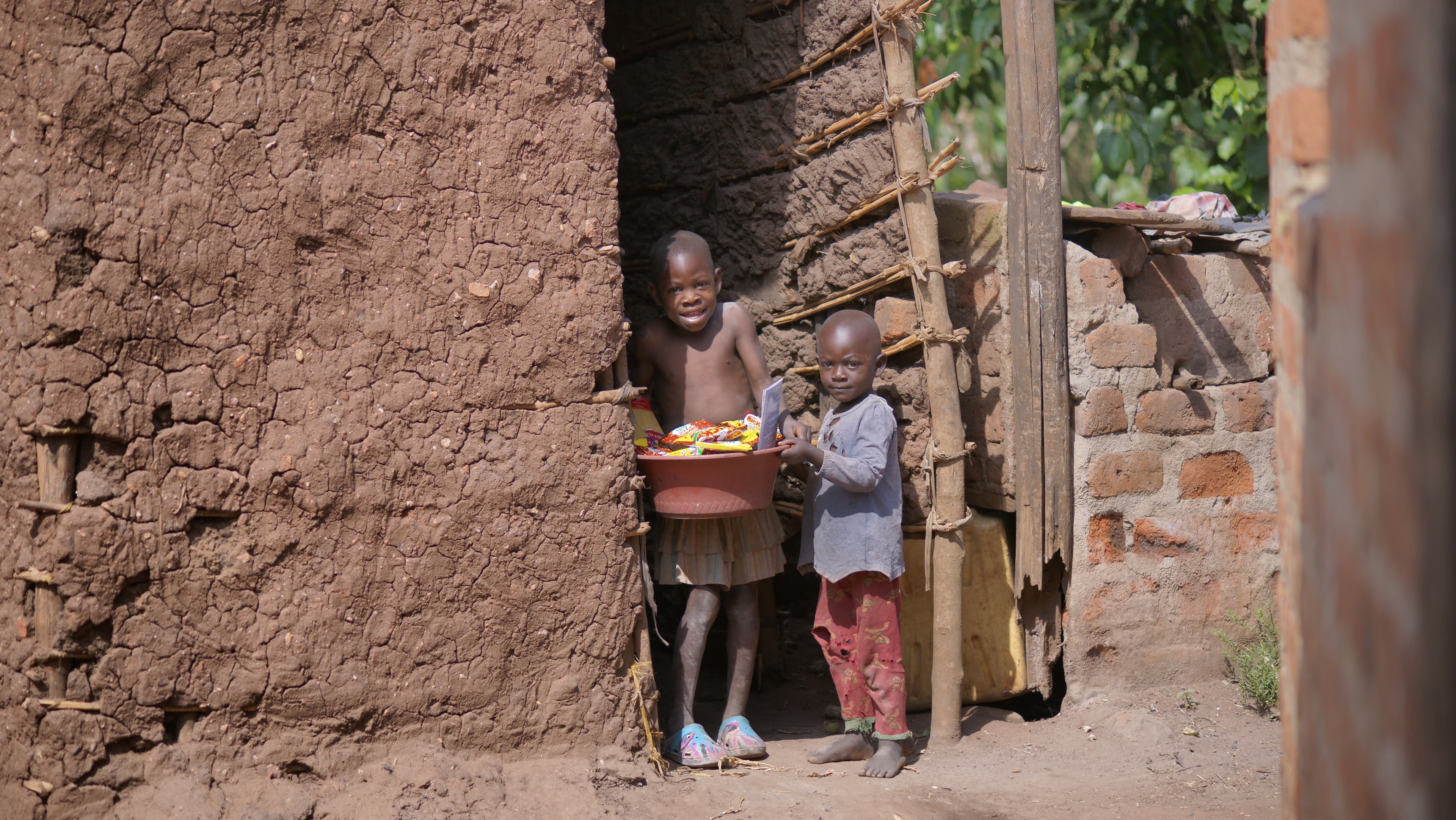 Two children stand at the entrance of a mud-brick house holding a basin of food.