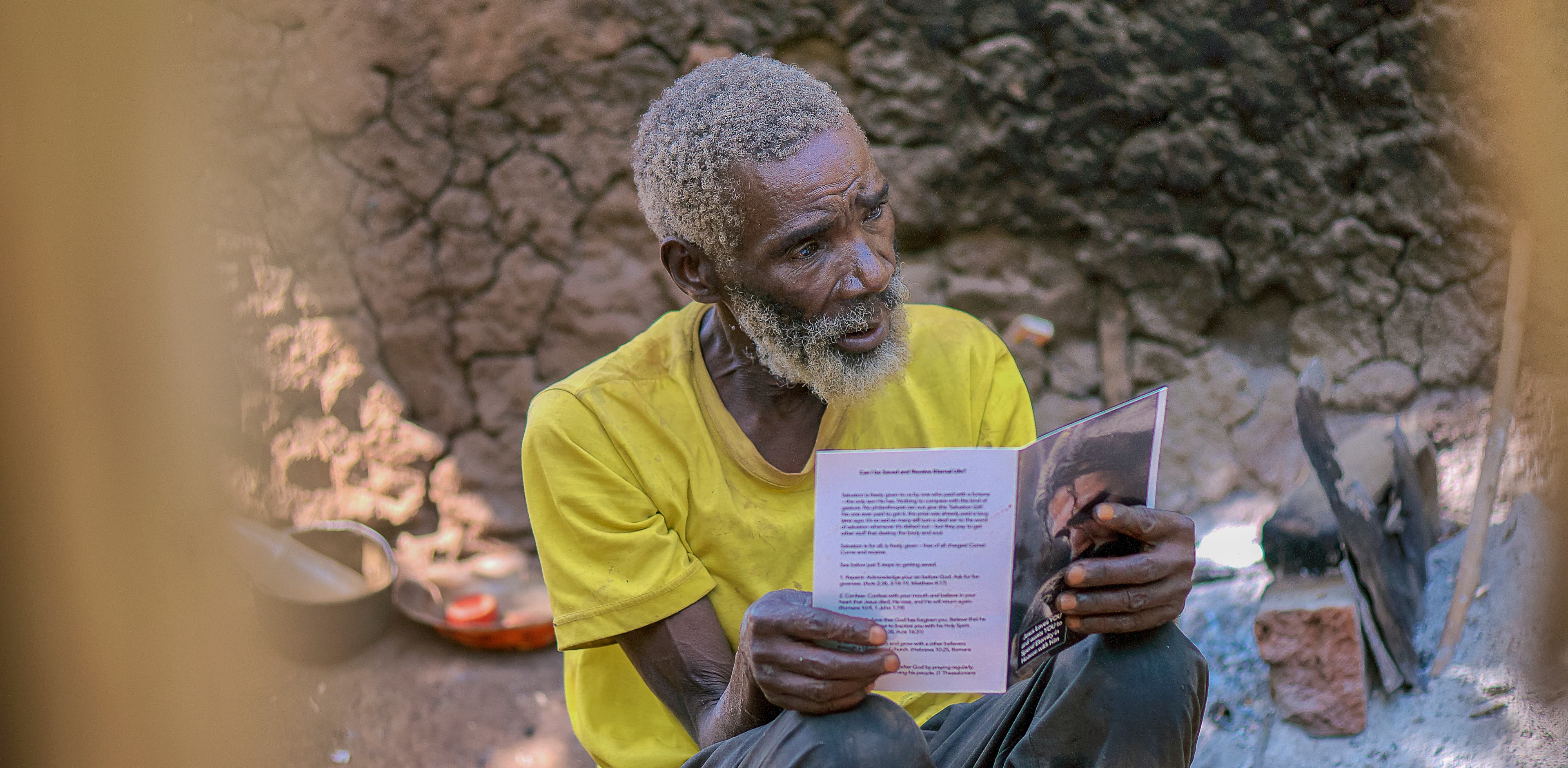 Close-up of an elderly man in a yellow shirt reading a paper tract in front of a brick wall.