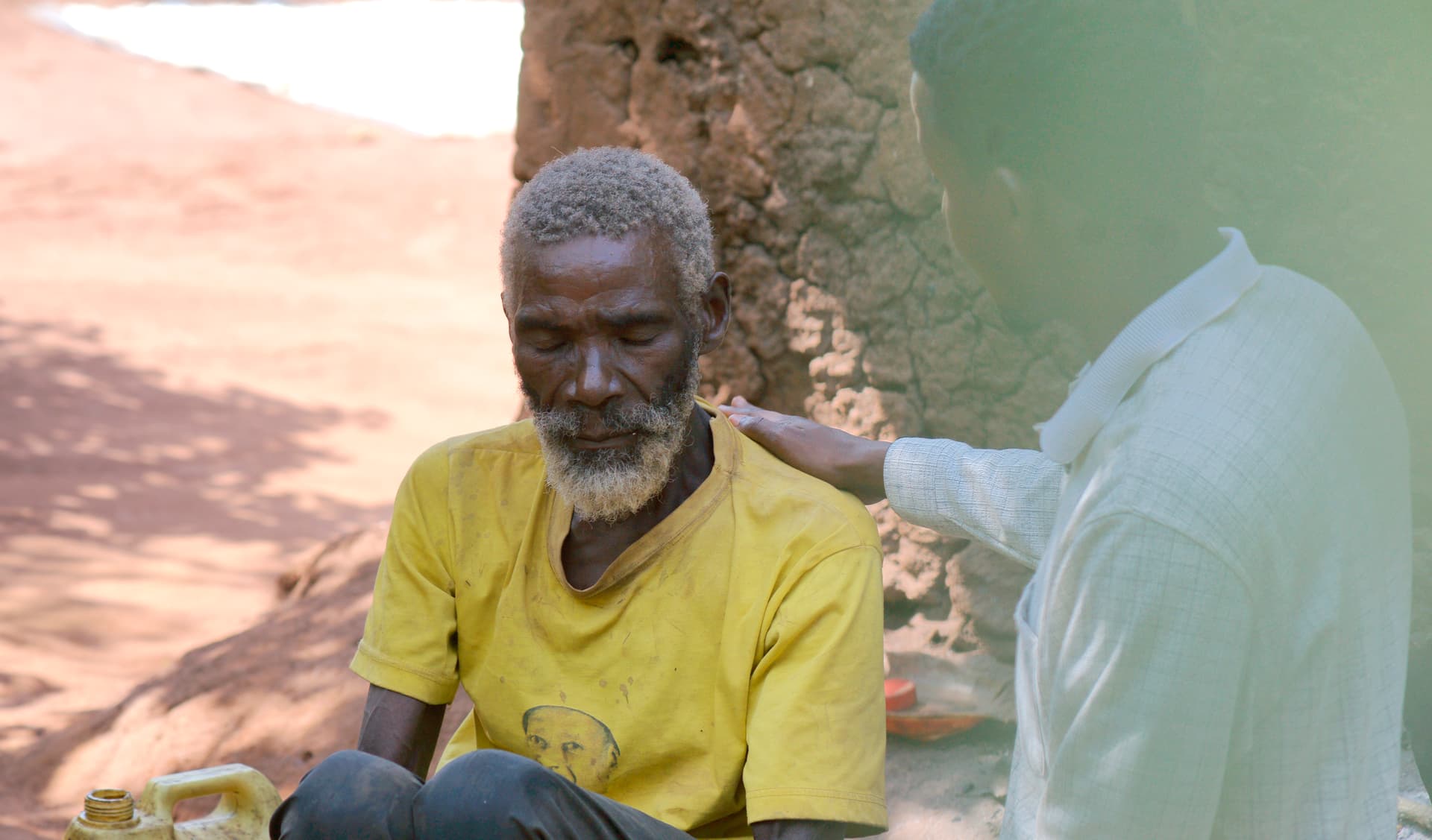Faded photo of an elderly man in a yellow shirt seated as someone reaches toward him outdoors.