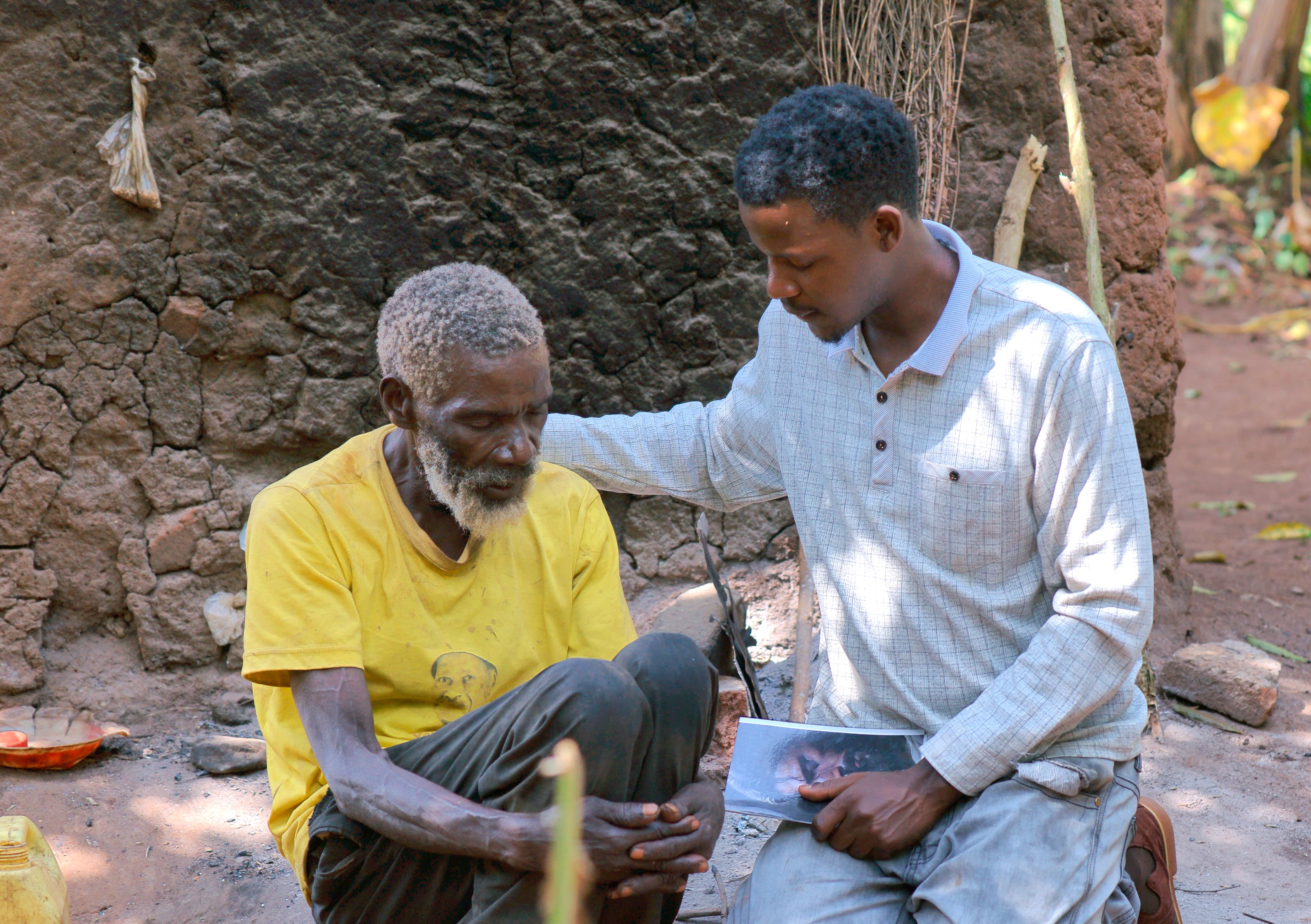 A JFM volunteer kneels beside an elderly man in a yellow shirt, handing him a paper outdoors.