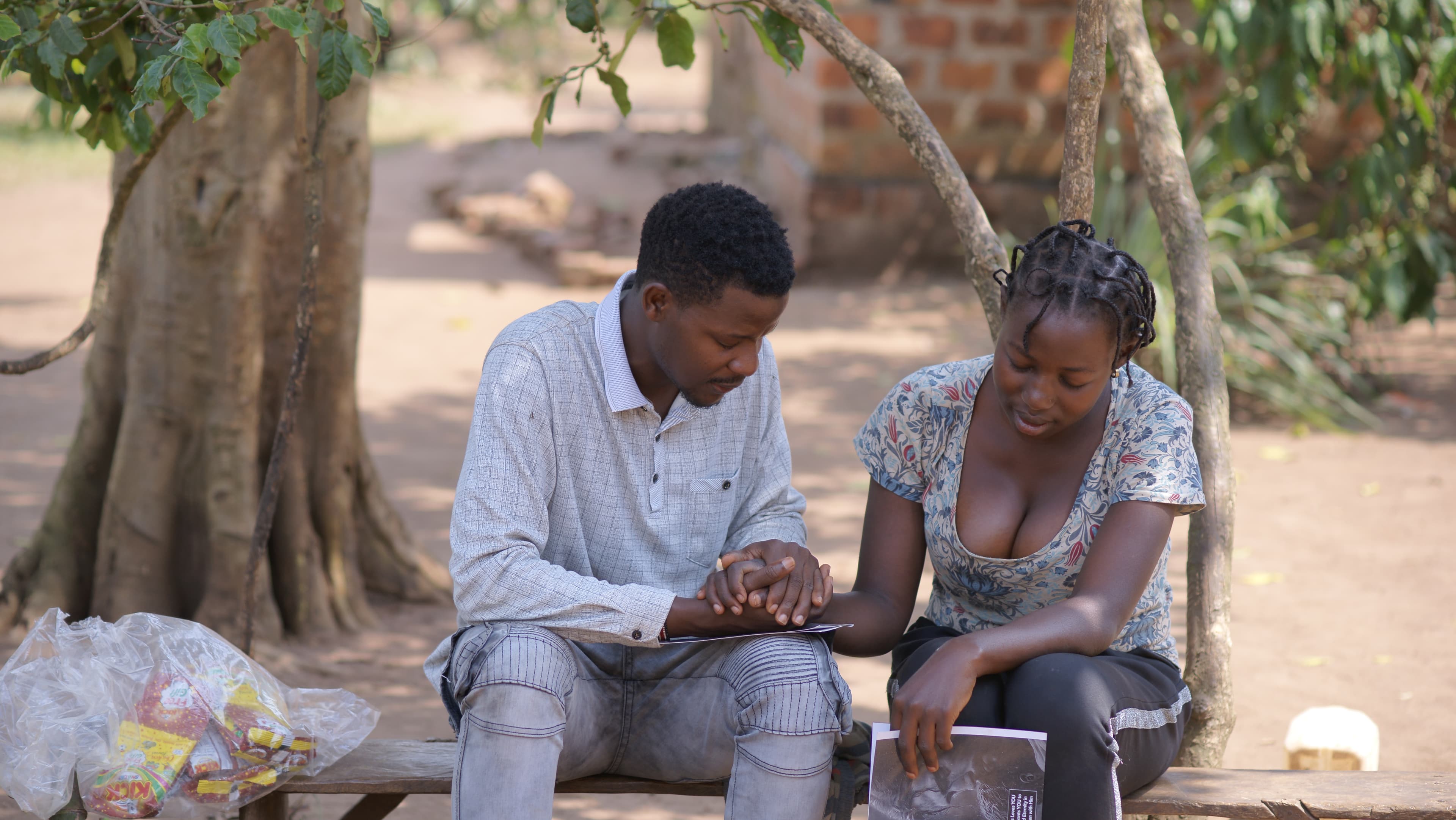 A young man and woman sit together under a tree, talking as they look over items.