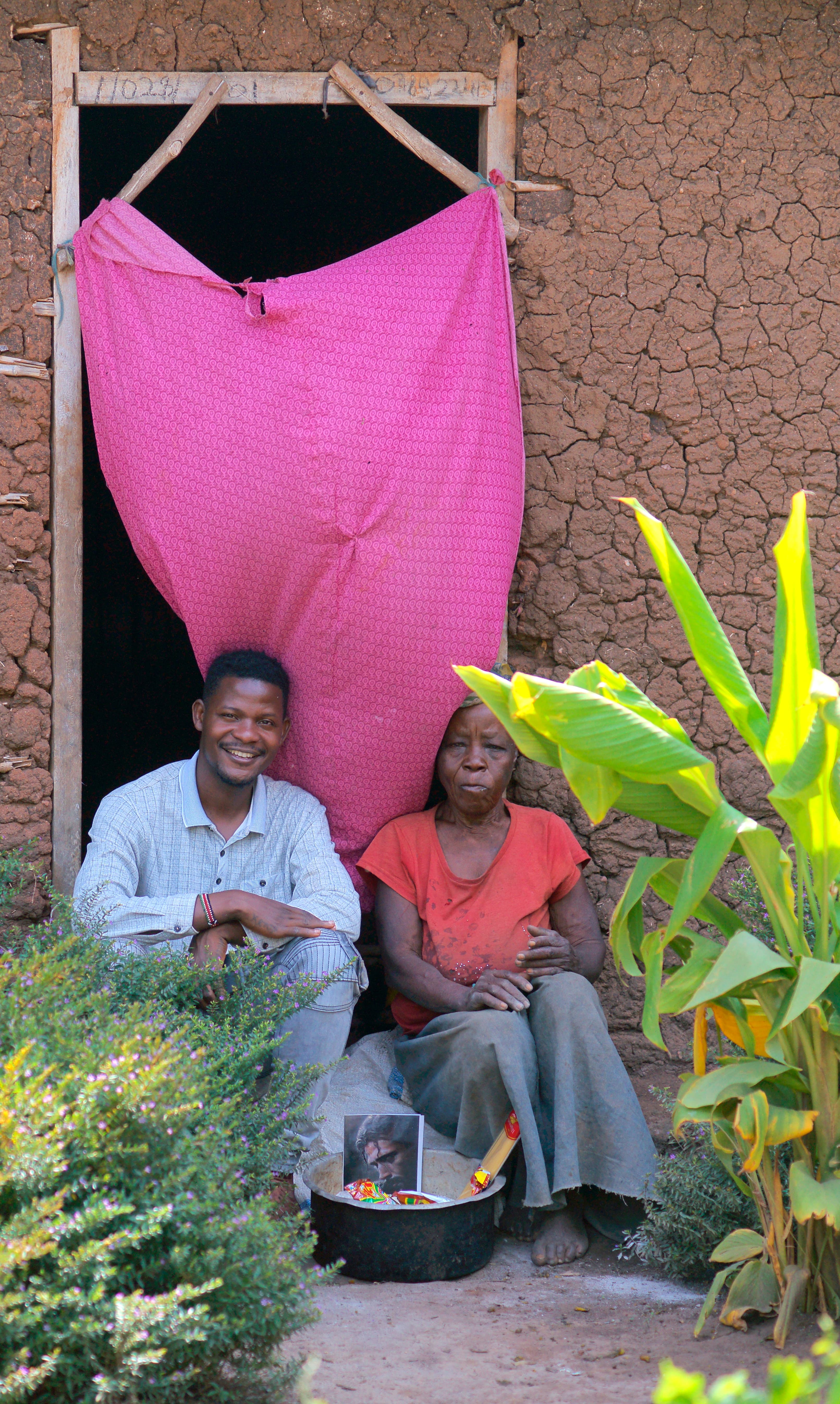 Two people sit in a doorway hung with a pink curtain, a food basket resting at their feet.