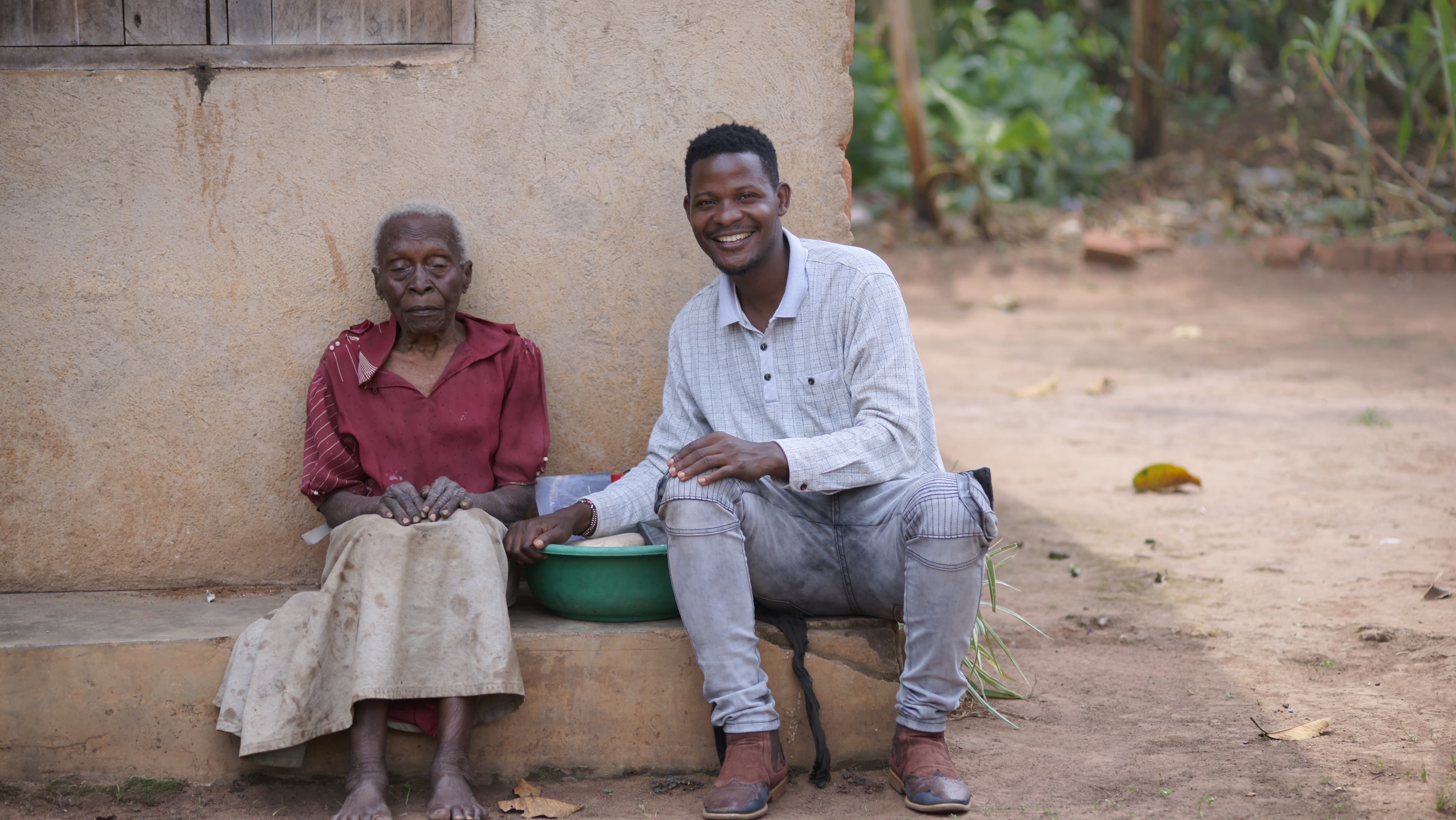 An elderly woman sits on the ground outside a mud house while a young man stands beside her.
