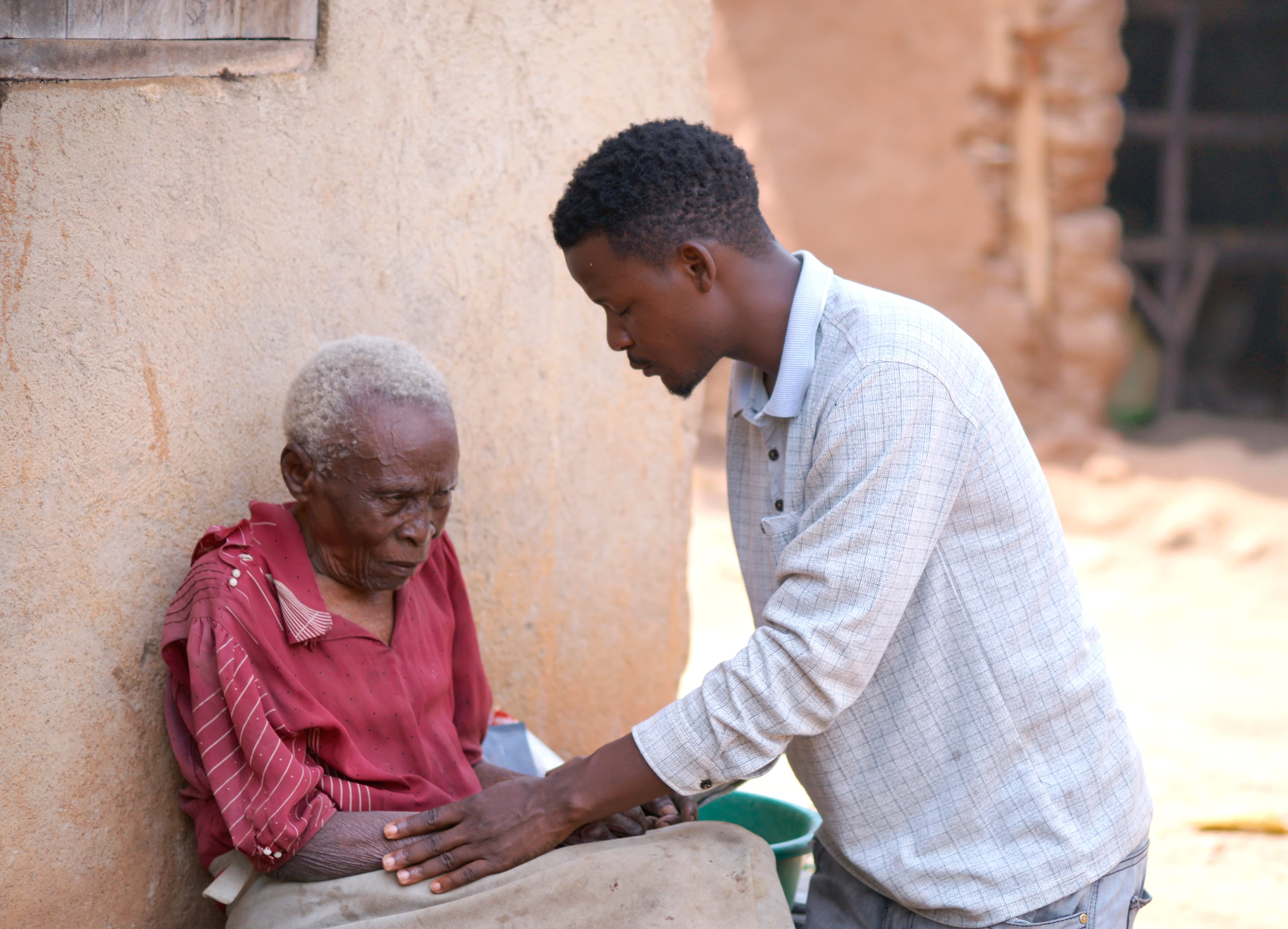 A young man kneels to help an elderly woman in red, holding a cup between them outdoors.