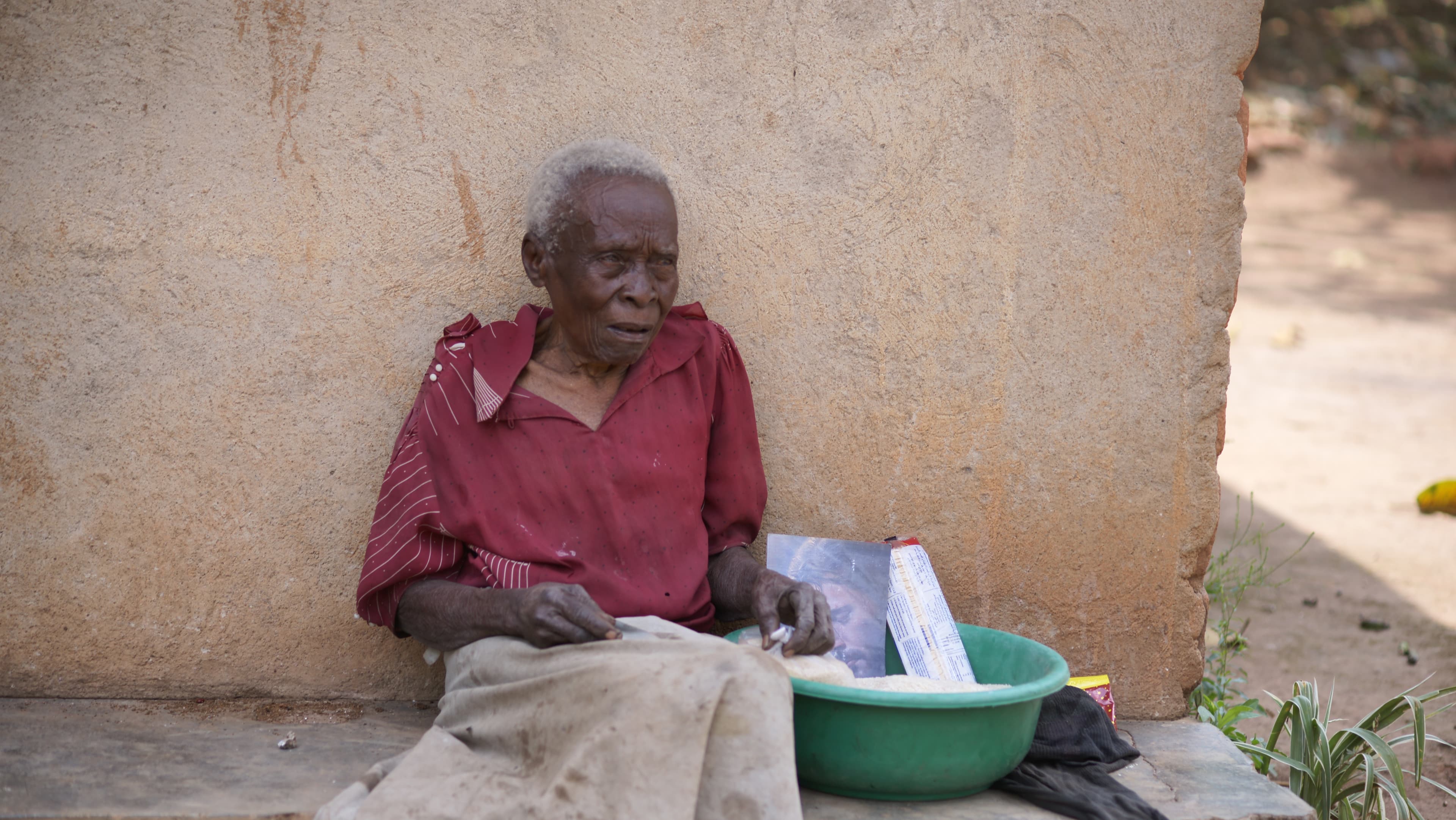An elderly woman in a red dress sits on the ground outdoors with a bowl and food items.