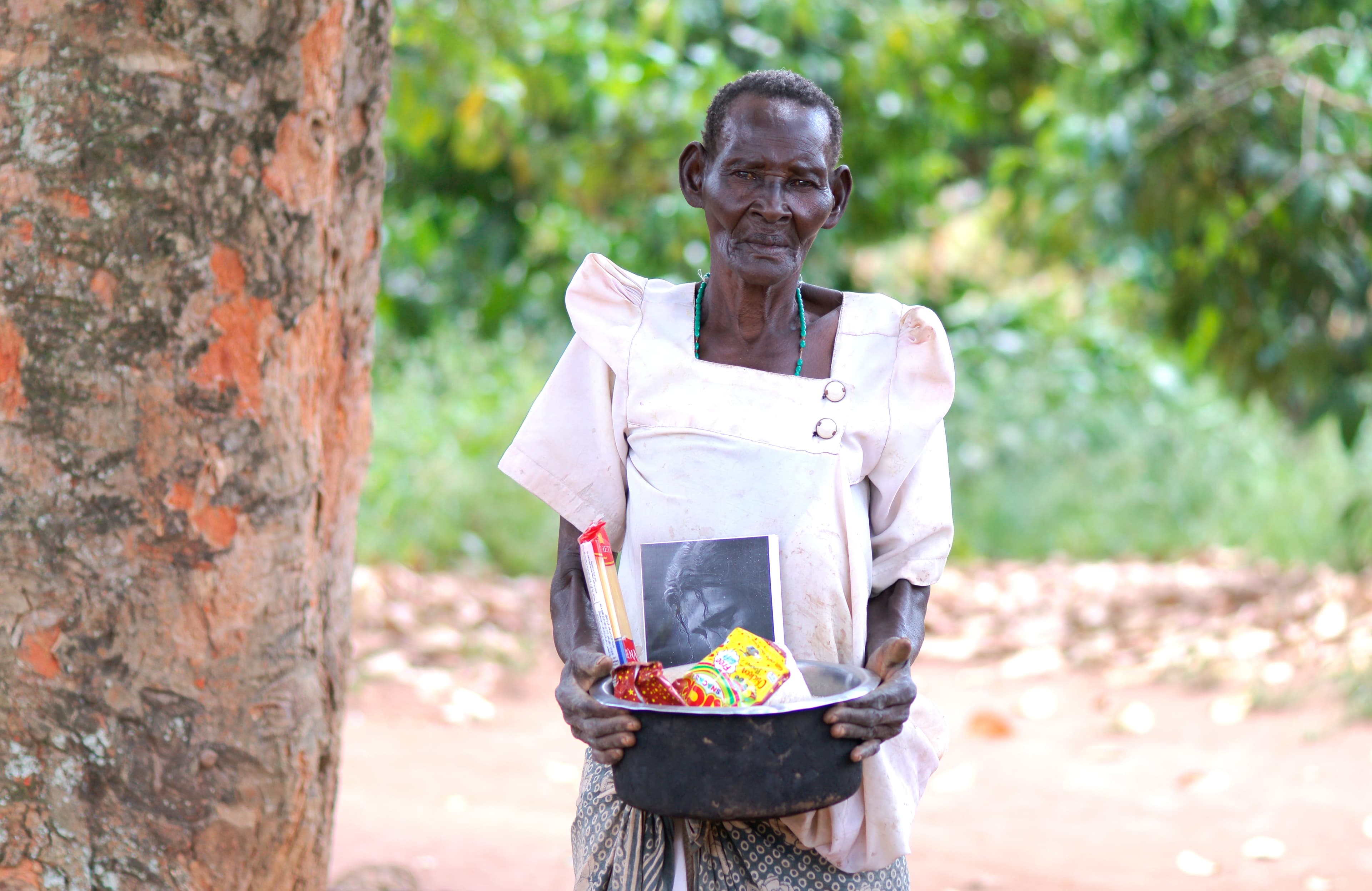 Elderly man standing outdoors holding a black basin of food items and papers
