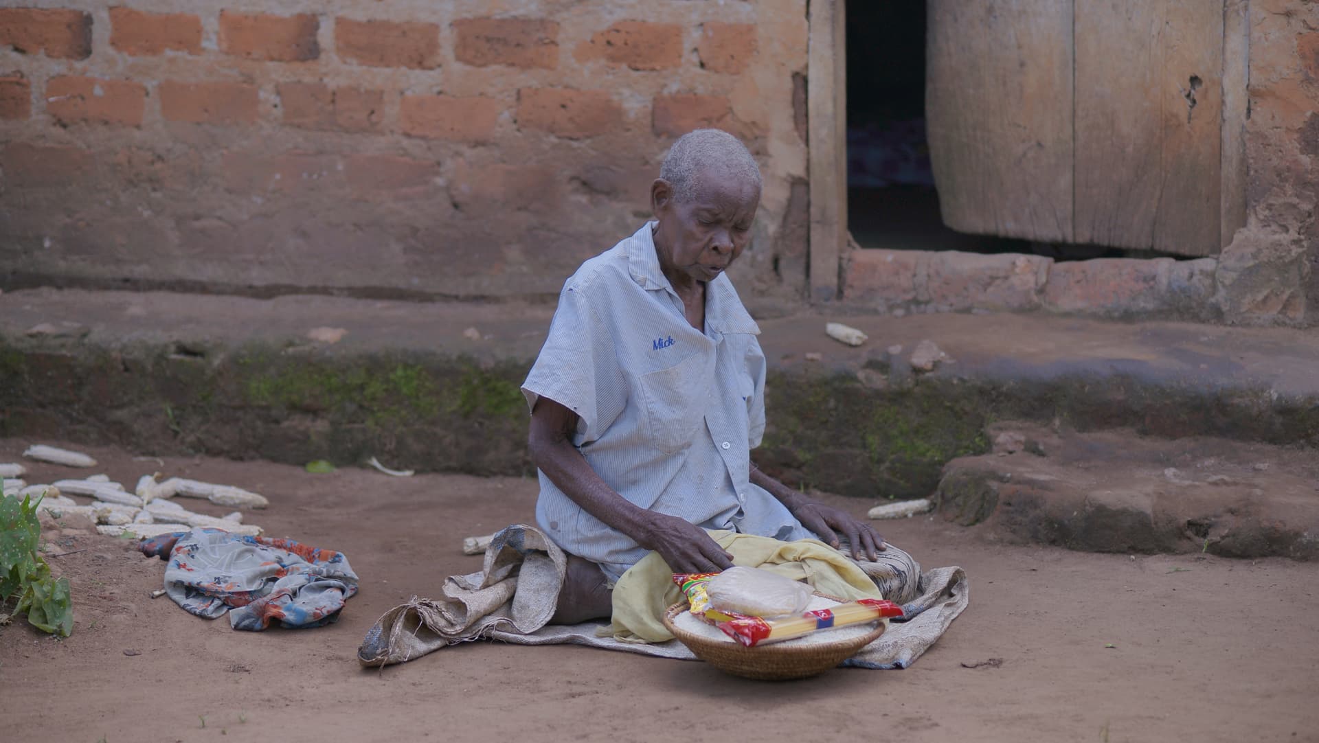An elderly woman kneels on the ground sorting food items laid out on a cloth.