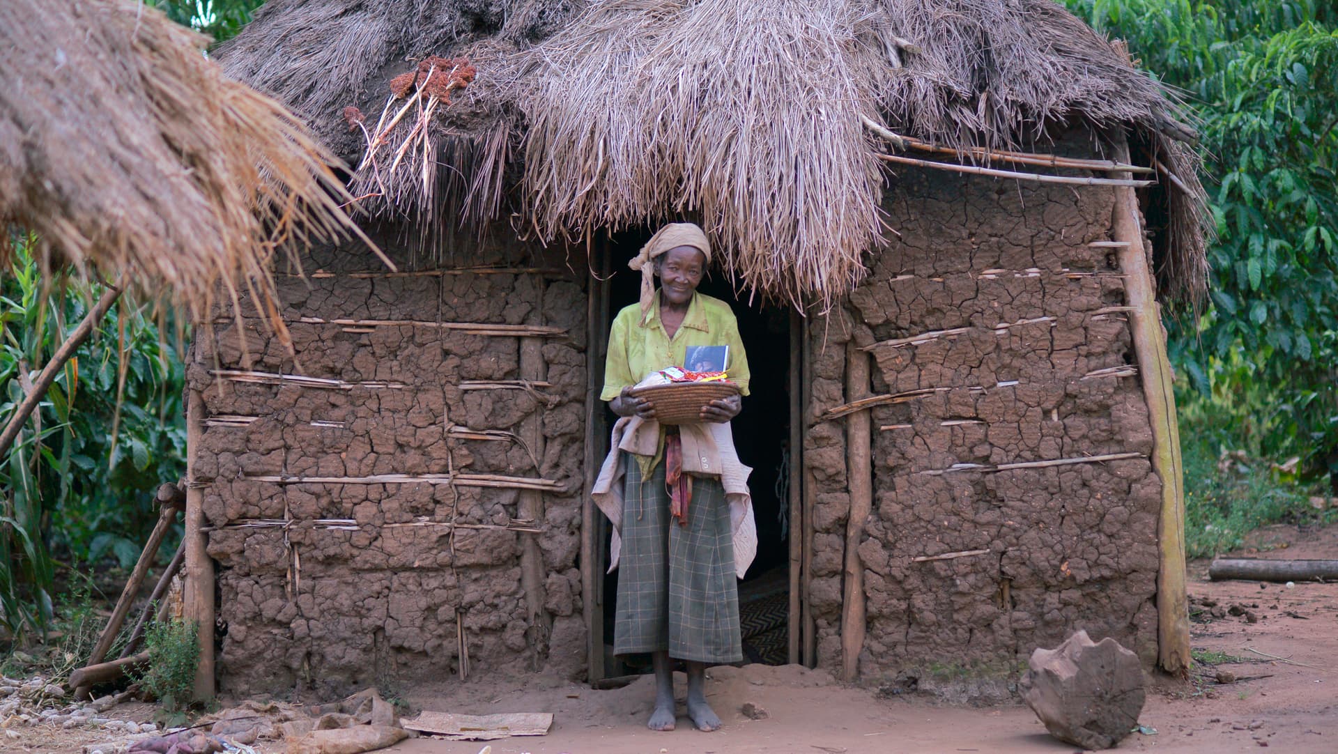 An elderly woman stands alone in the doorway of a thatched-roof mud hut.