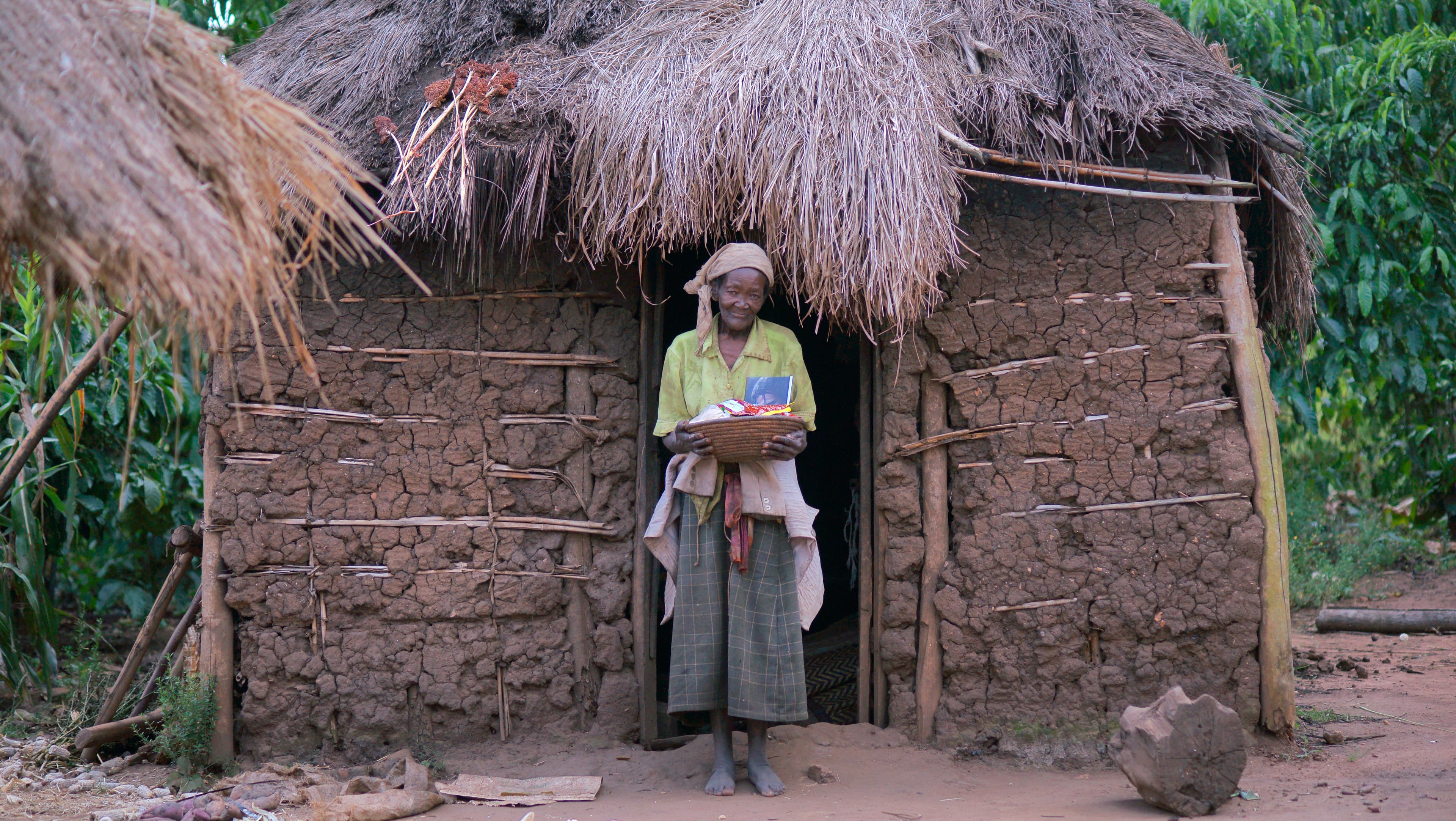 An elderly woman stands alone in the doorway of a thatched-roof mud hut.