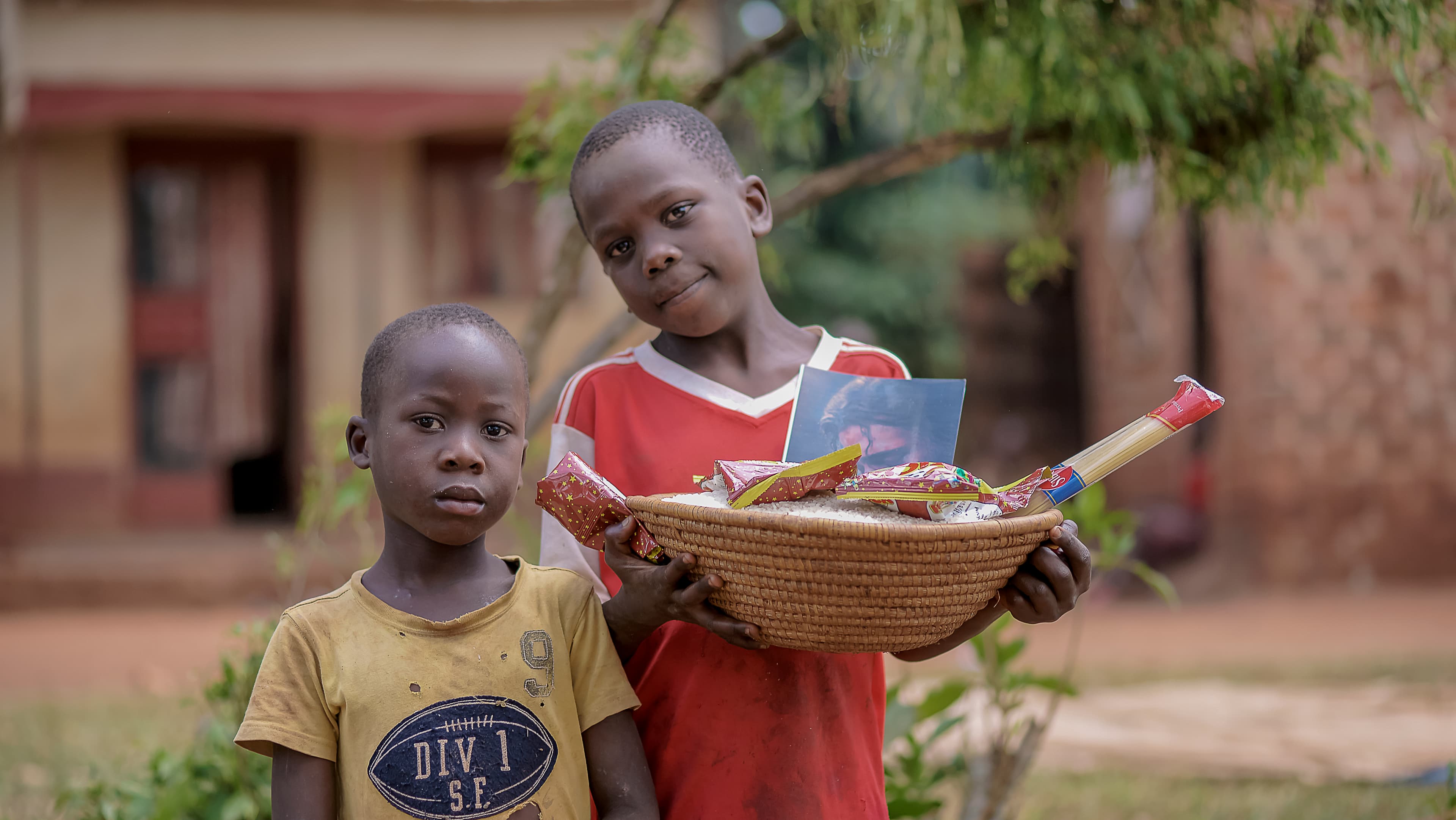 Two children standing together outdoors, one holding a wicker basket of food
