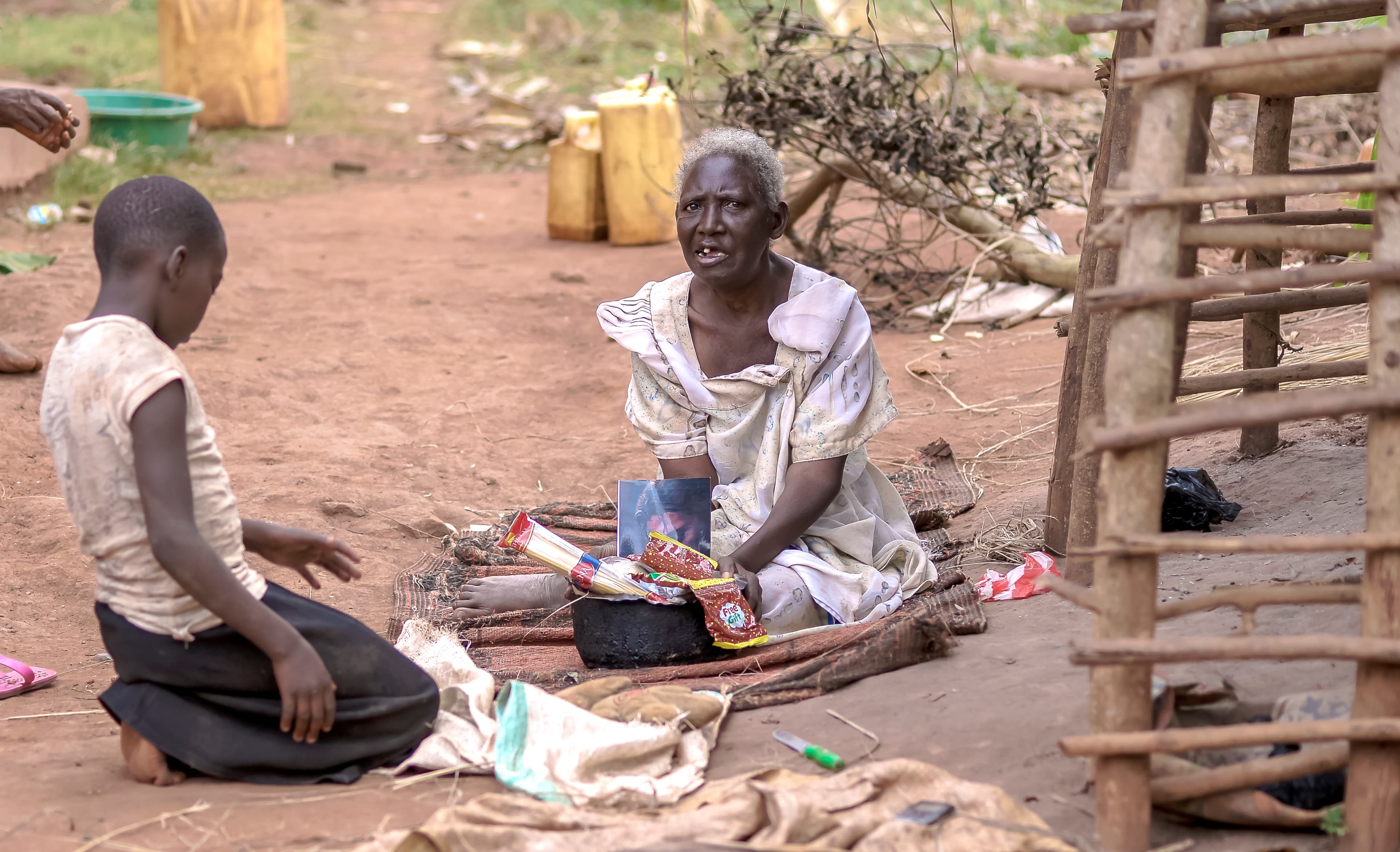 Elderly woman and a child sitting outdoors near a woven fence with yellow jerry cans