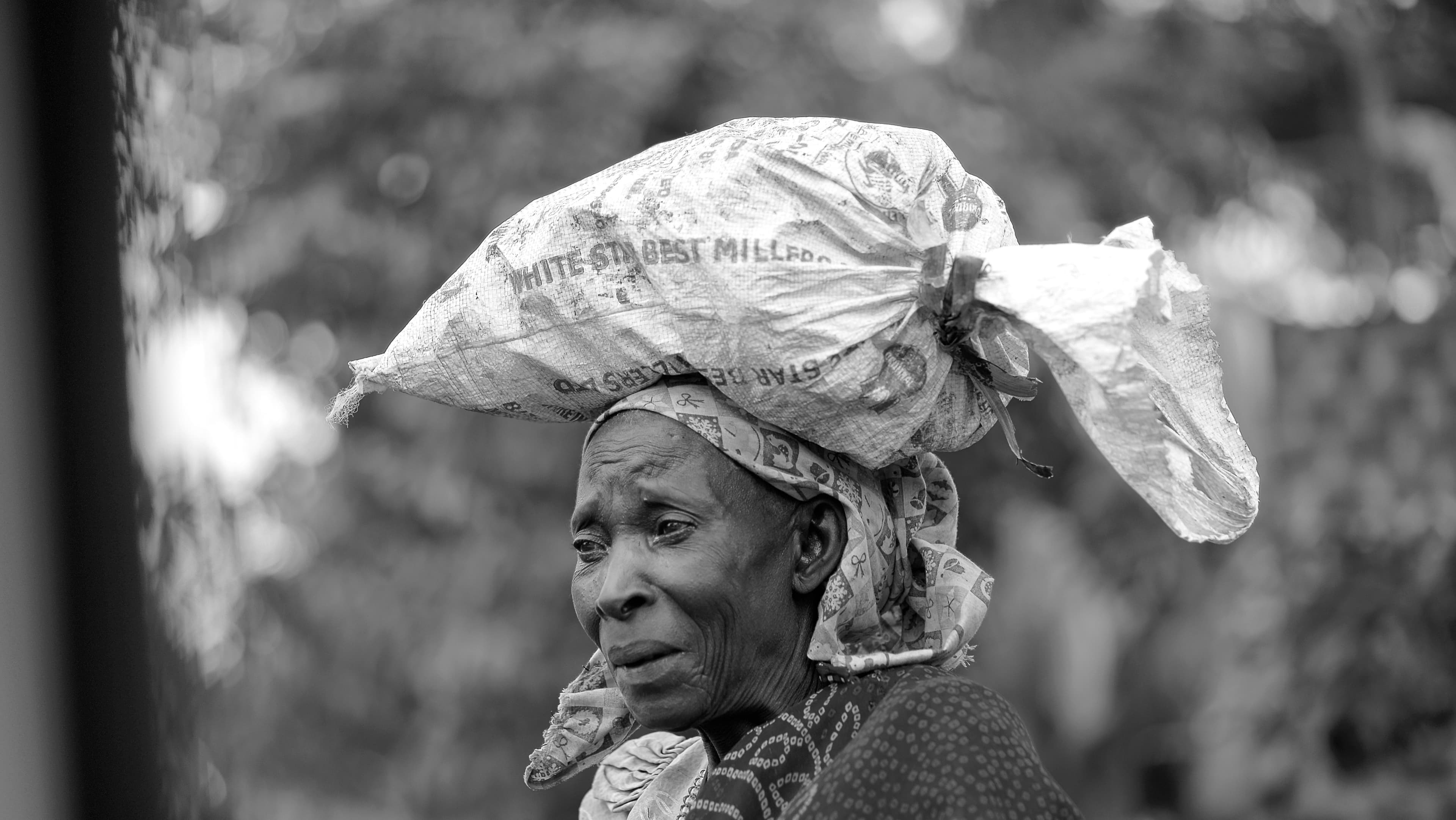 Black and white photo of an elderly woman carrying a bundle on her head