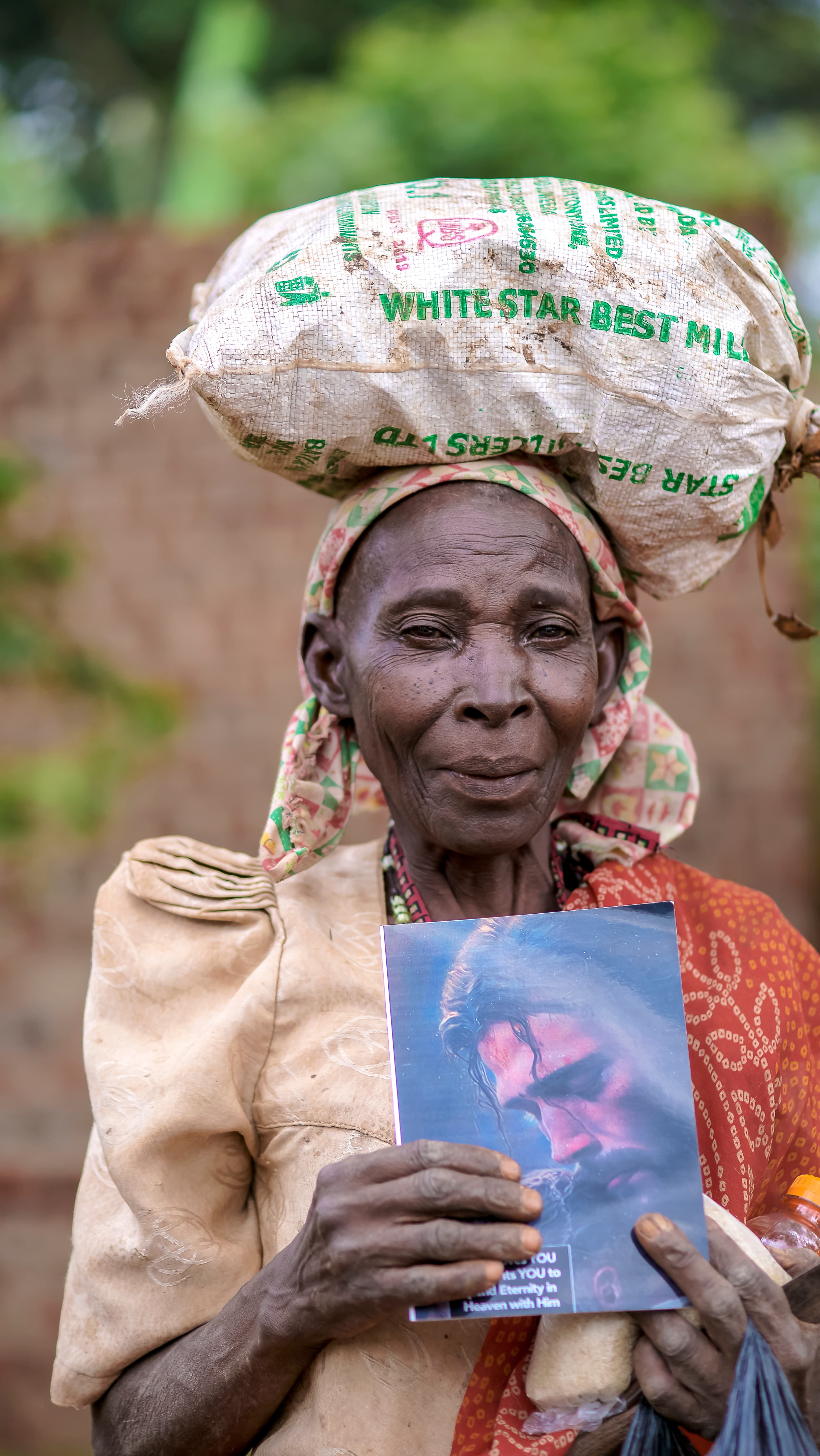 Elderly woman carrying a white sack of maize flour on her head while holding a photo card