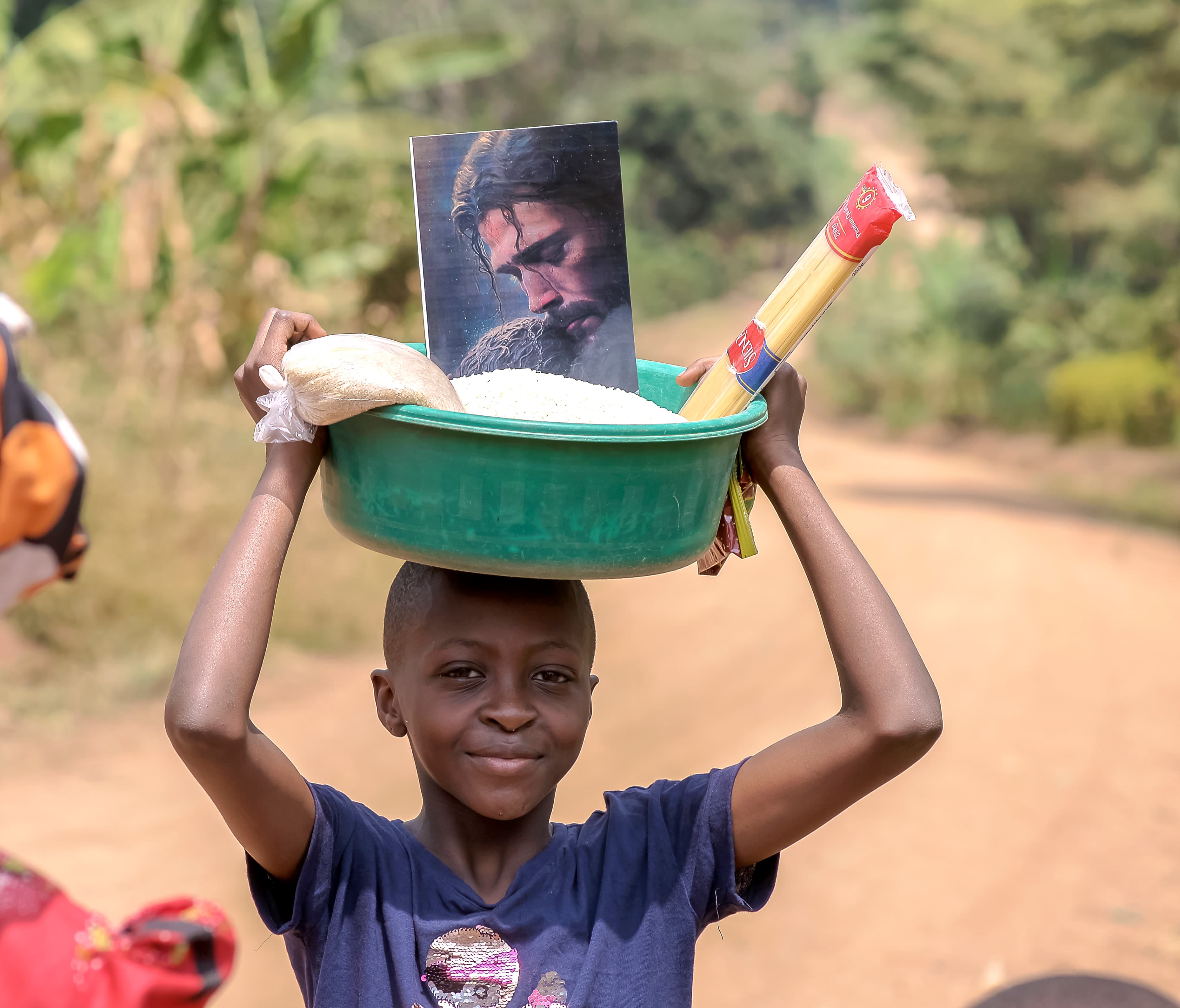 Boy carrying a green basin with food and a photo card on his head while walking along a dirt road