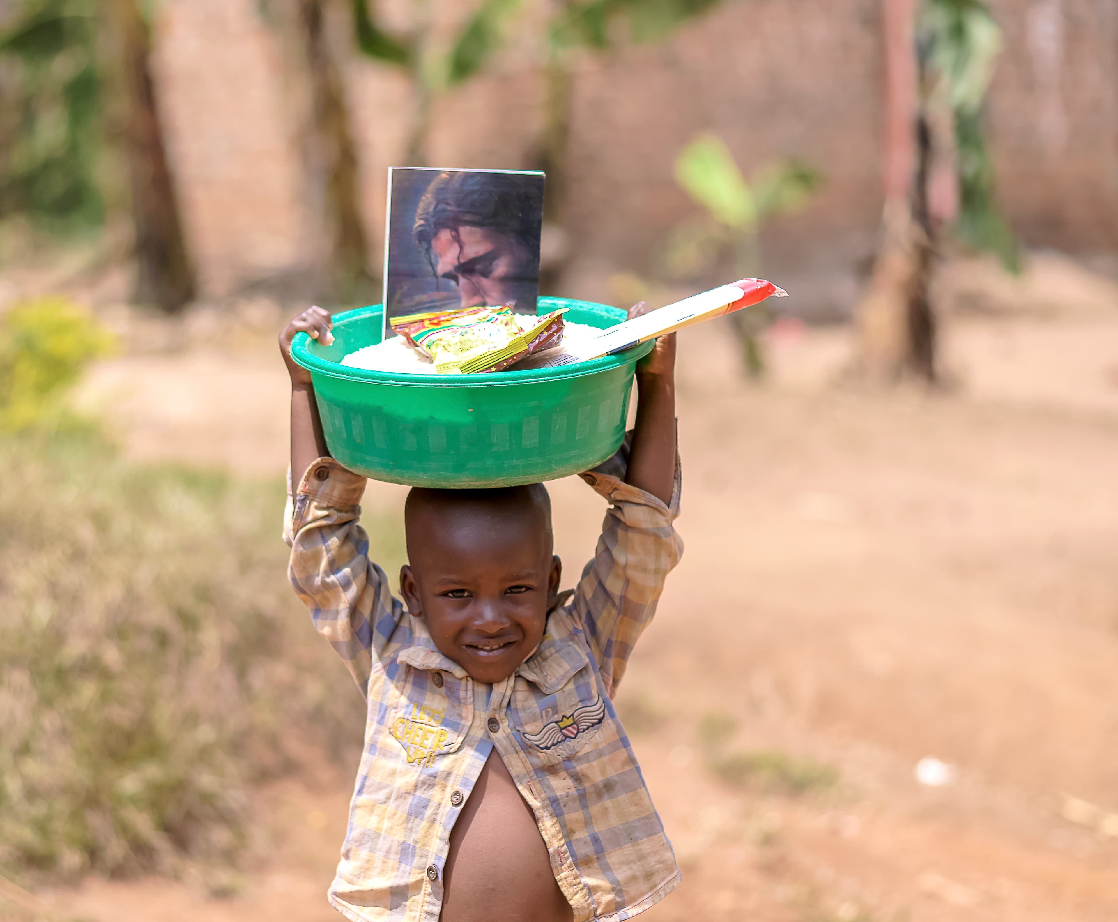 Boy carrying a green basin with food and a photo card balanced on his head outdoors