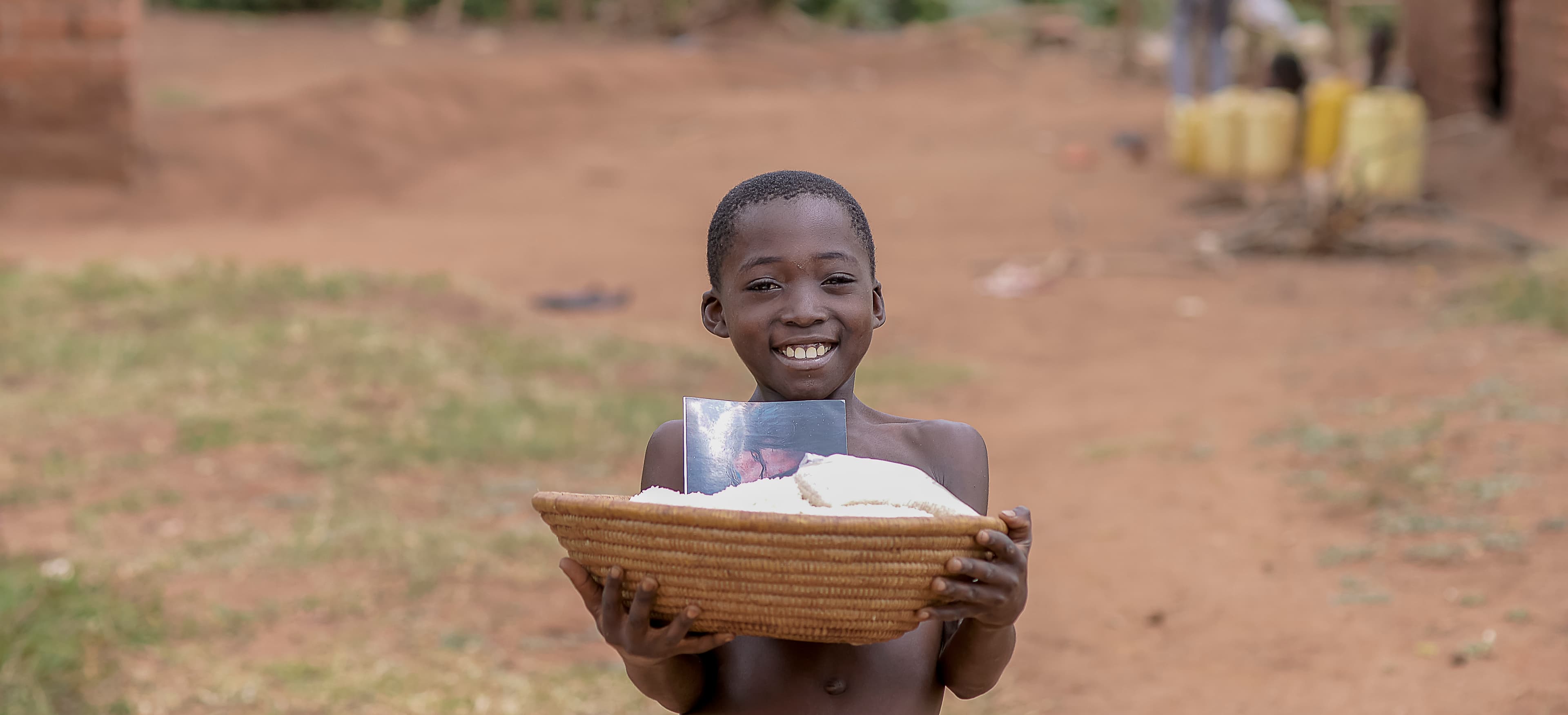 Smiling boy holding a woven basket with bread and food items, standing on bare dirt ground