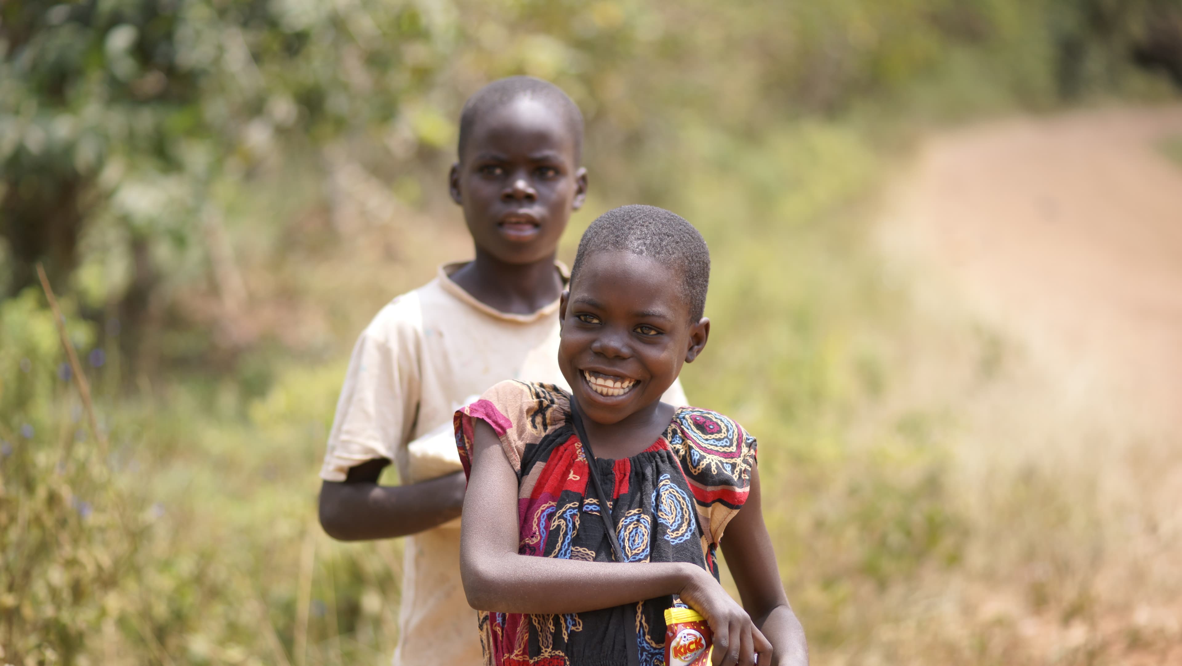 Two children standing together outdoors on a dirt path, one holding a photo card