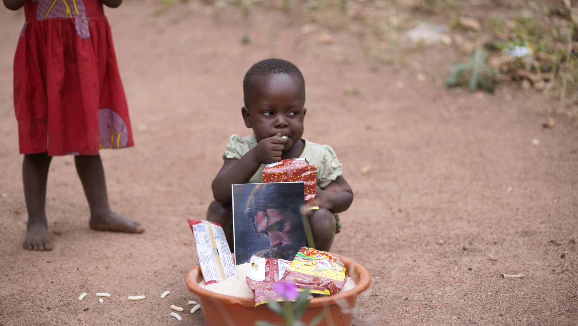 Young child in a red dress standing beside a basket of food gifts and a photo card on the ground