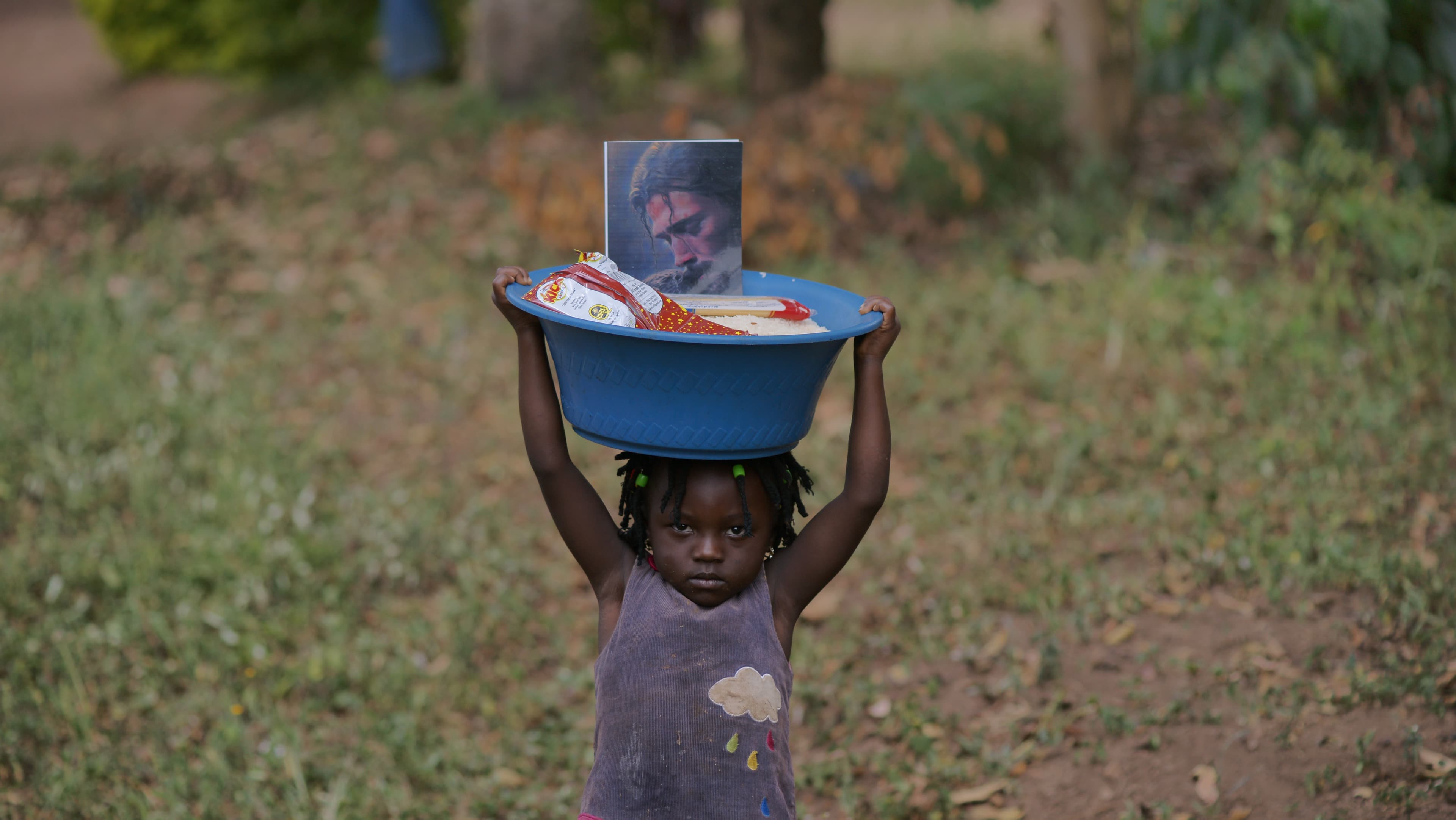 Child carrying a basin topped with a photo card on their head outdoors