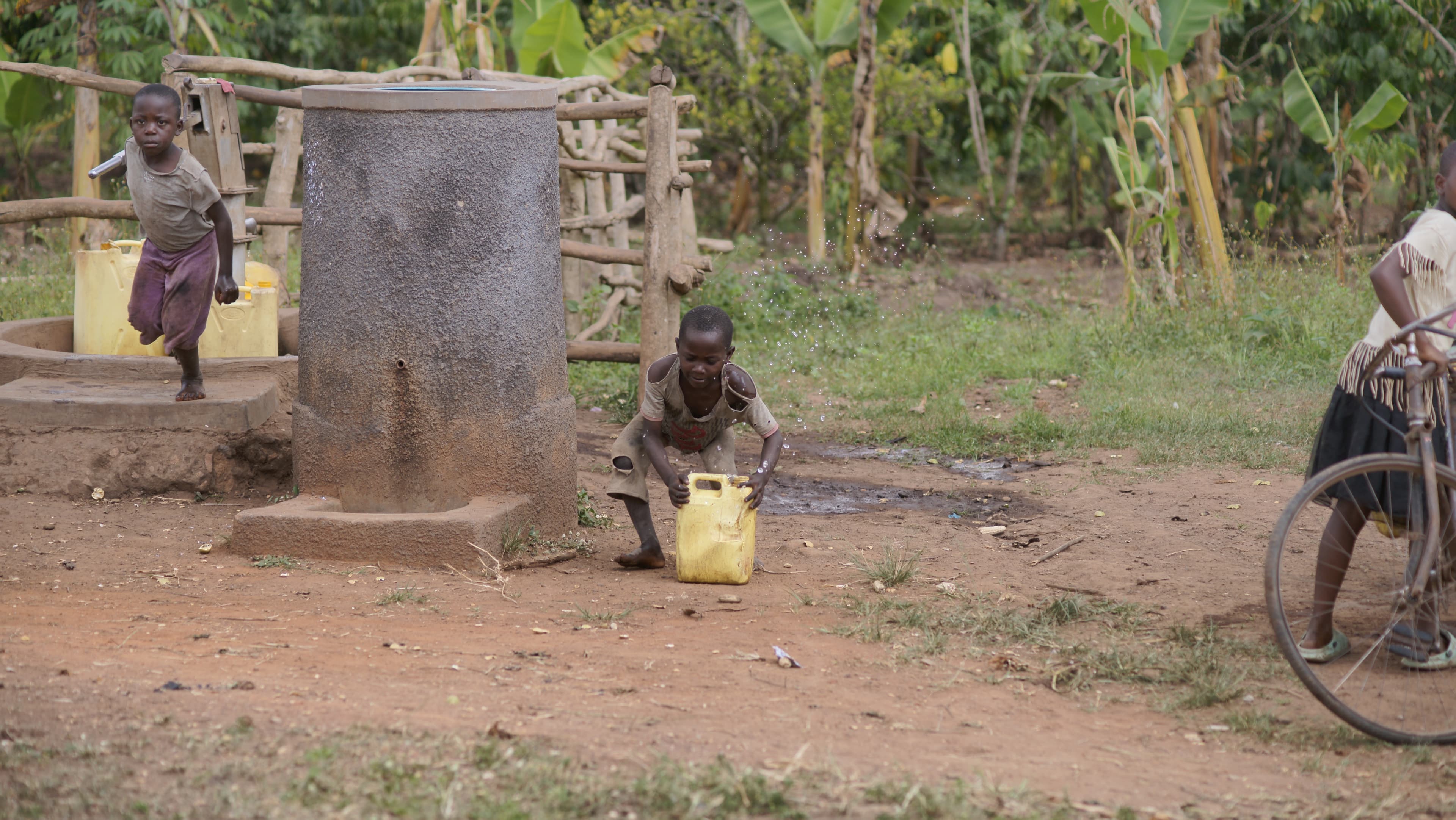 Child at a rural water pump station with another child on a bicycle nearby