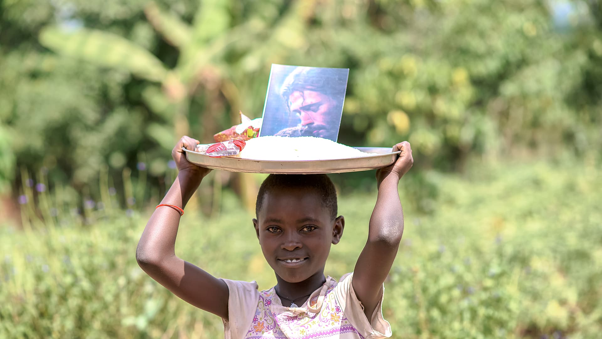 Child carrying a white basin topped with a photo card balanced on their head outdoors
