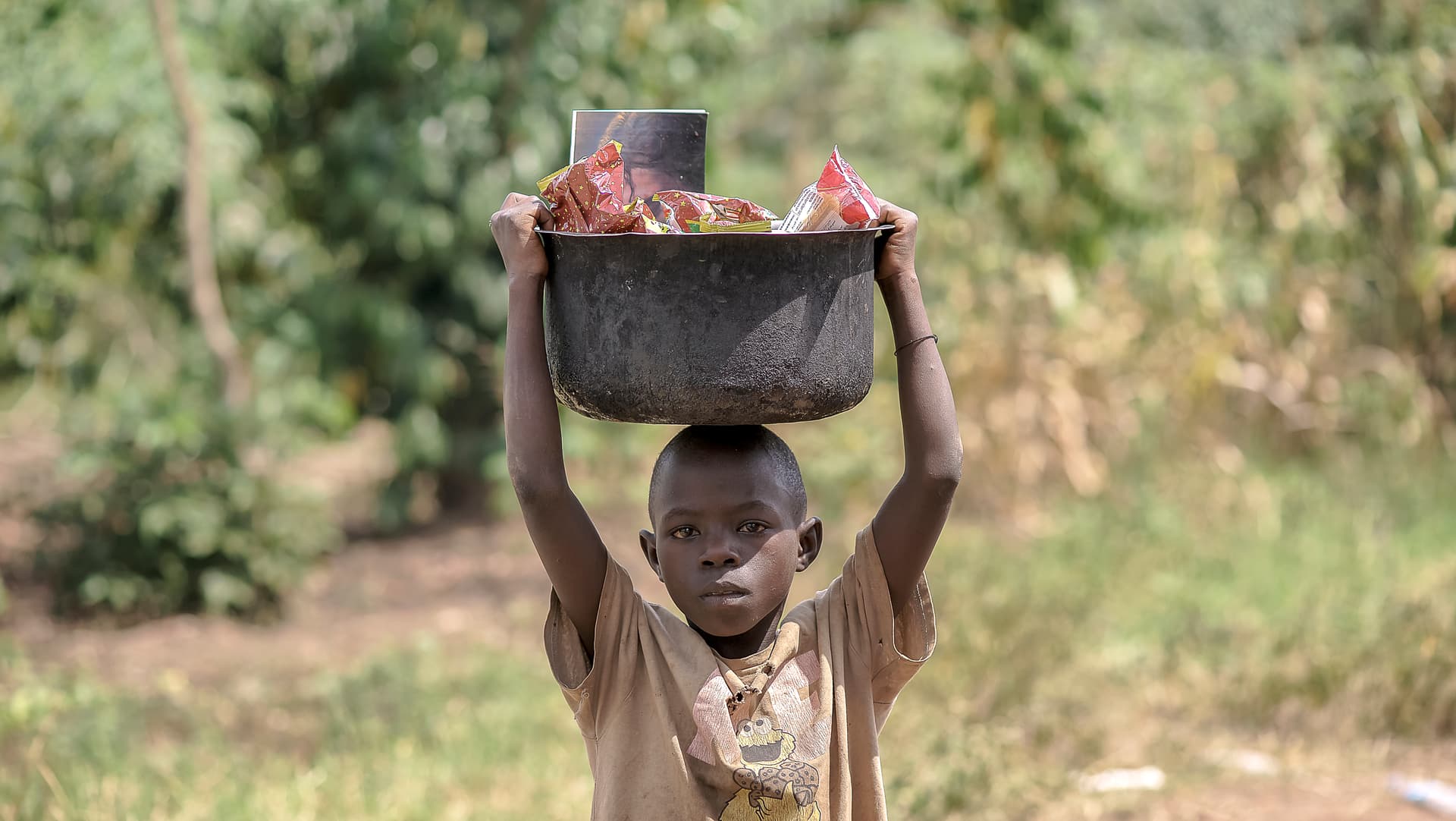 Child carrying a dark basin topped with a photo card on their head while walking outdoors