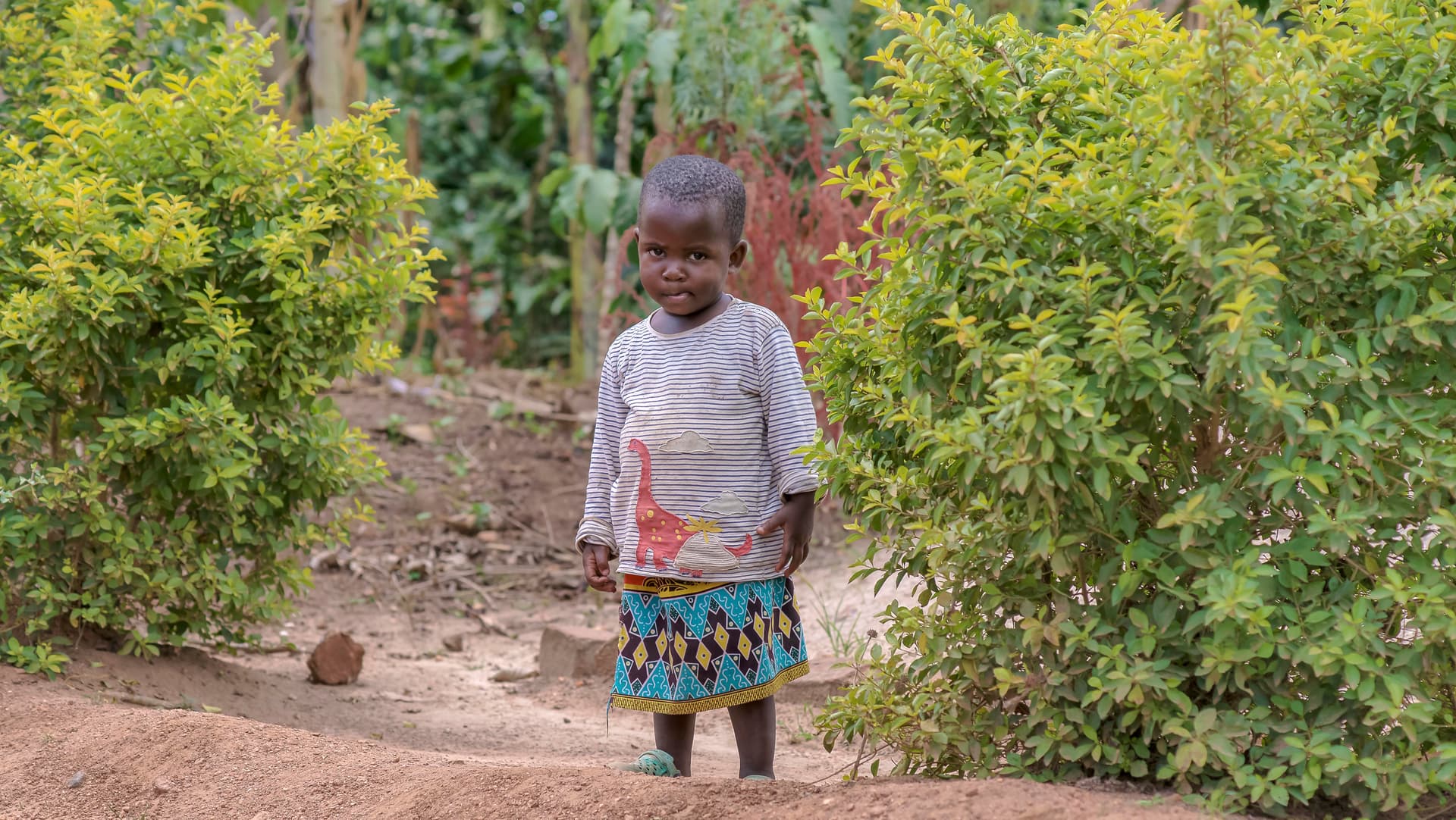 Child standing alone on a dirt path near green bushes in a Ugandan village