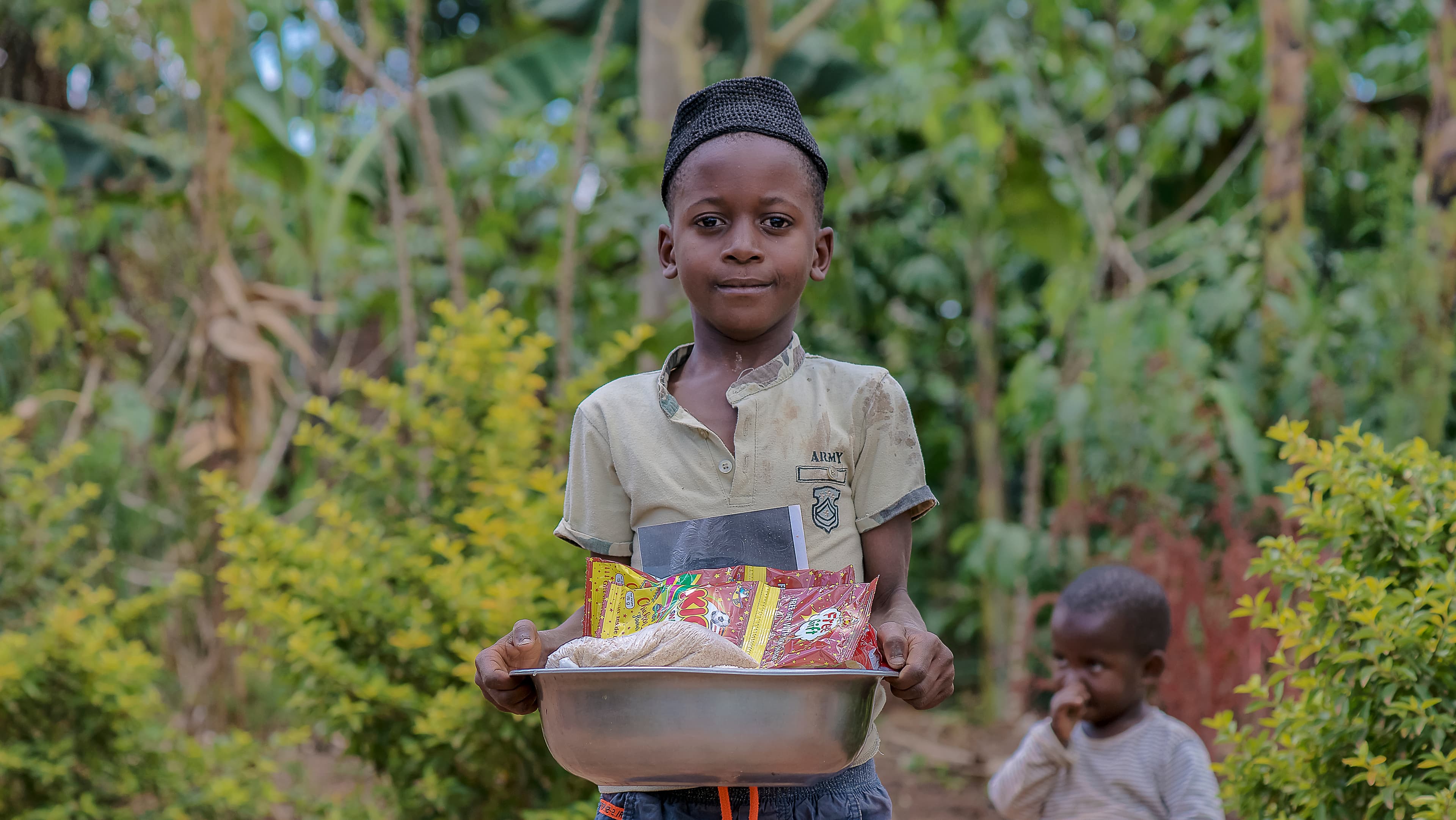 Child carrying a bowl of food supplies on their head while walking a rural path with another child