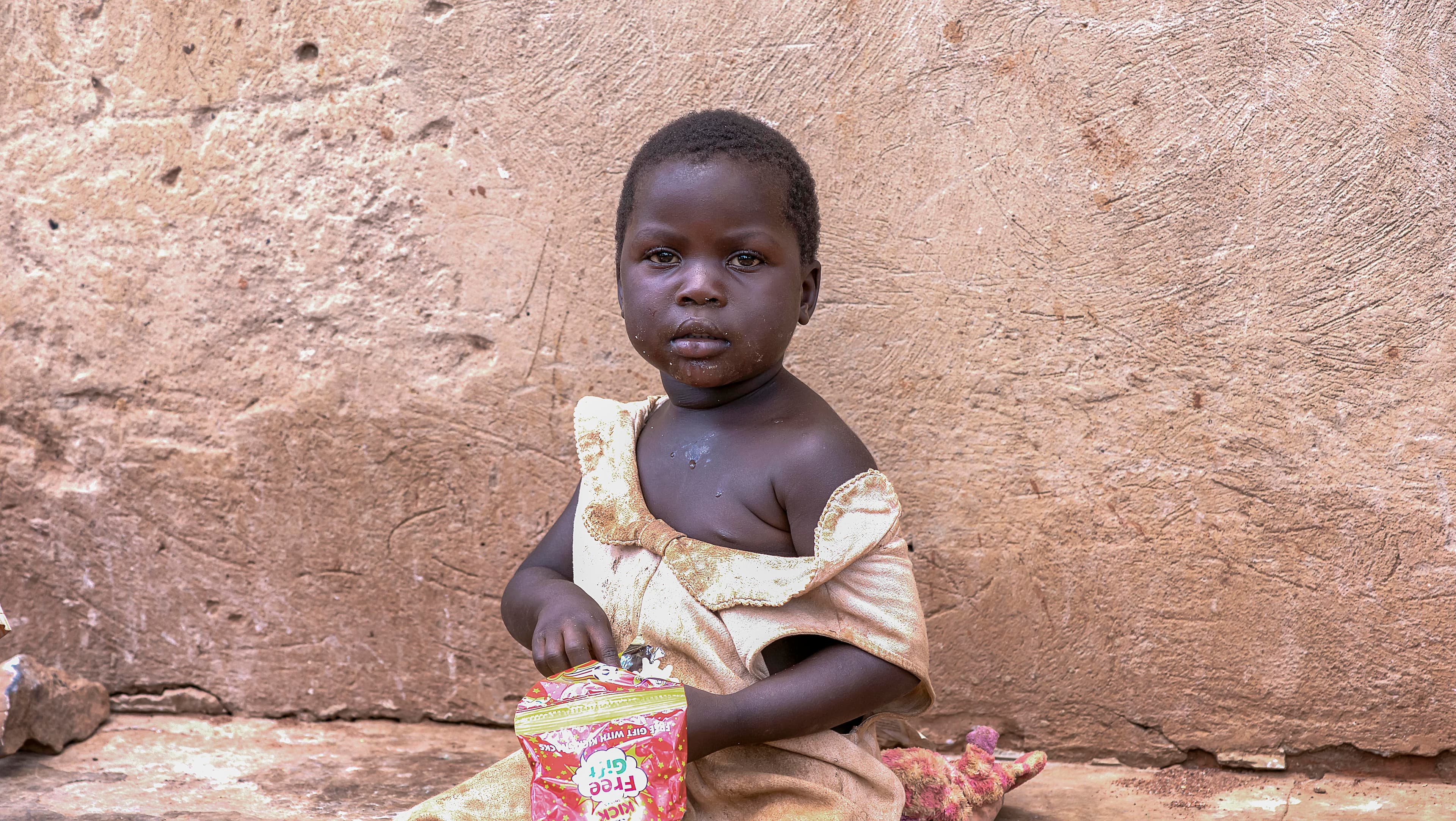 Young child sitting alone against a mud-brick wall wearing a light-colored dress