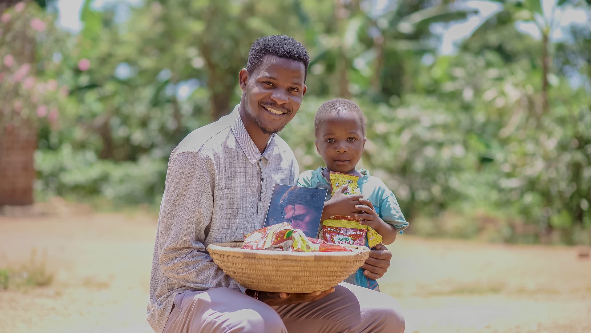 Man crouching outdoors beside a young child, holding a basket of food items
