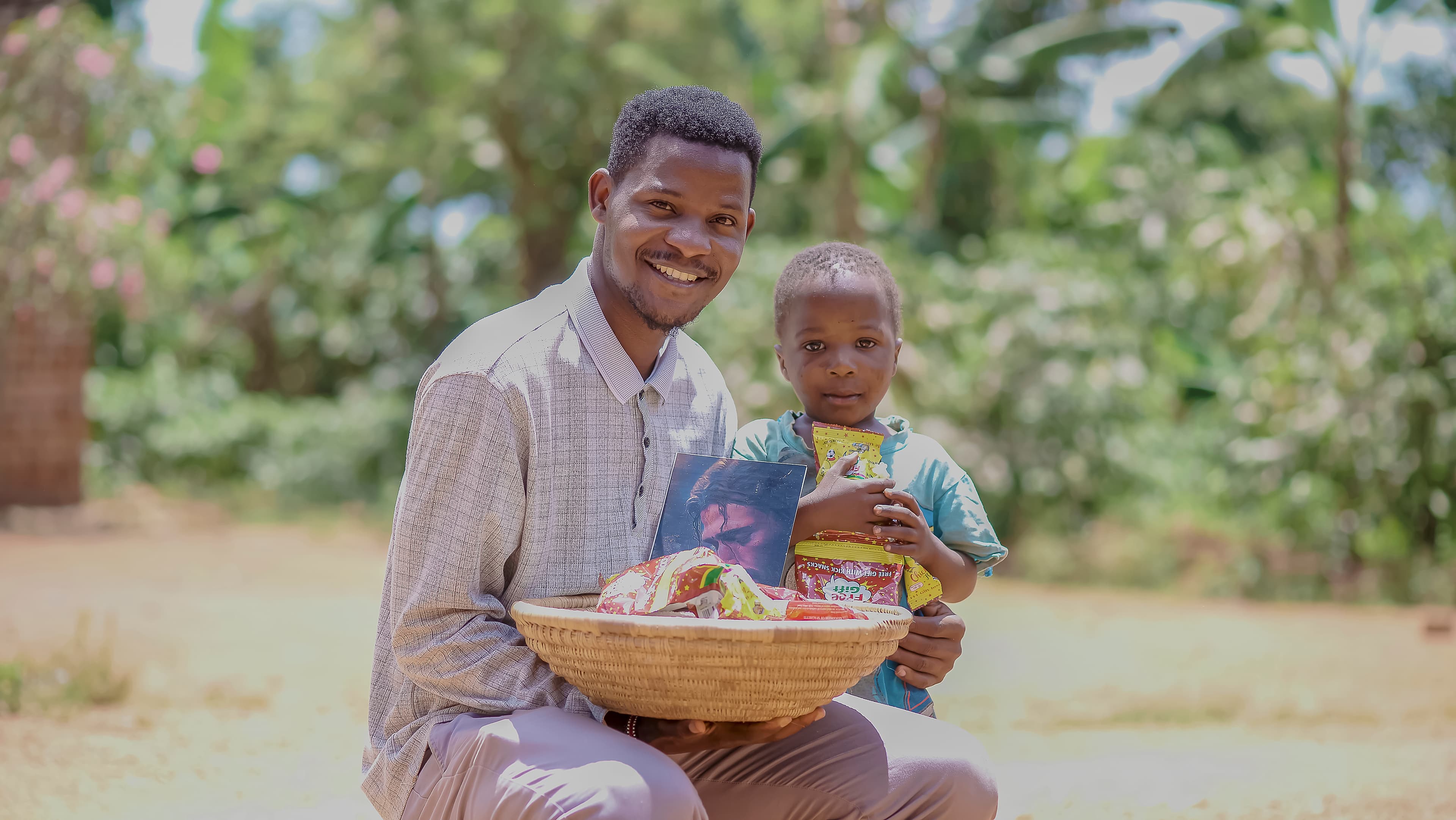 Man crouching outdoors beside a young child, holding a basket of food items