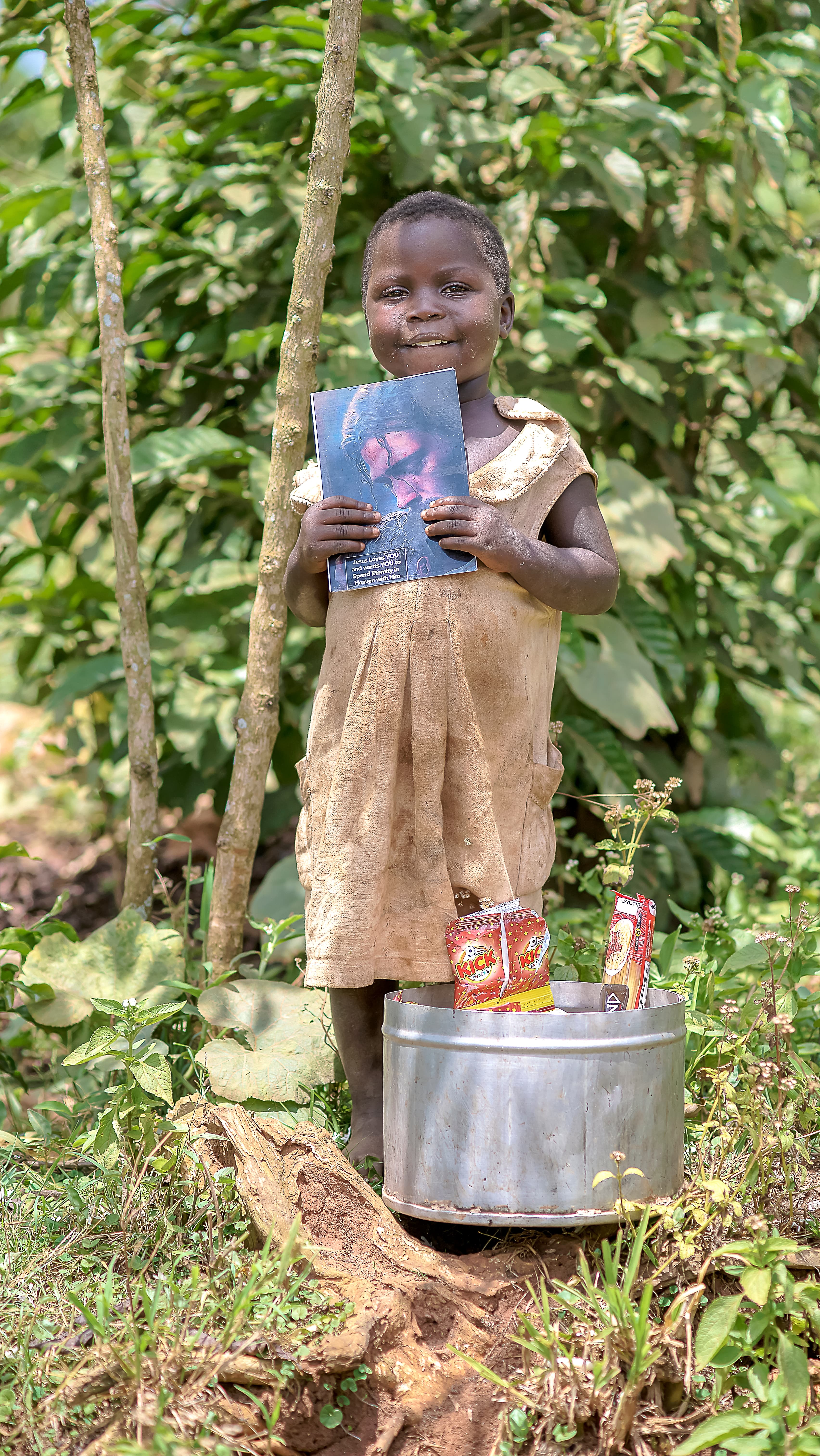 Girl standing among green plants holding a photo card and small items, with a tin basin at her feet