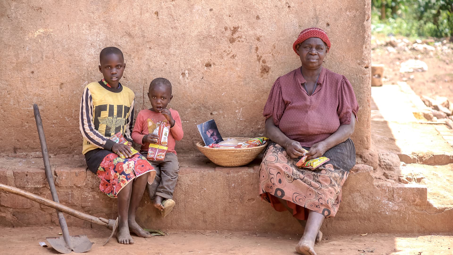 Elderly woman and children sitting outside a mud home with a food bowl and a walking stick nearby