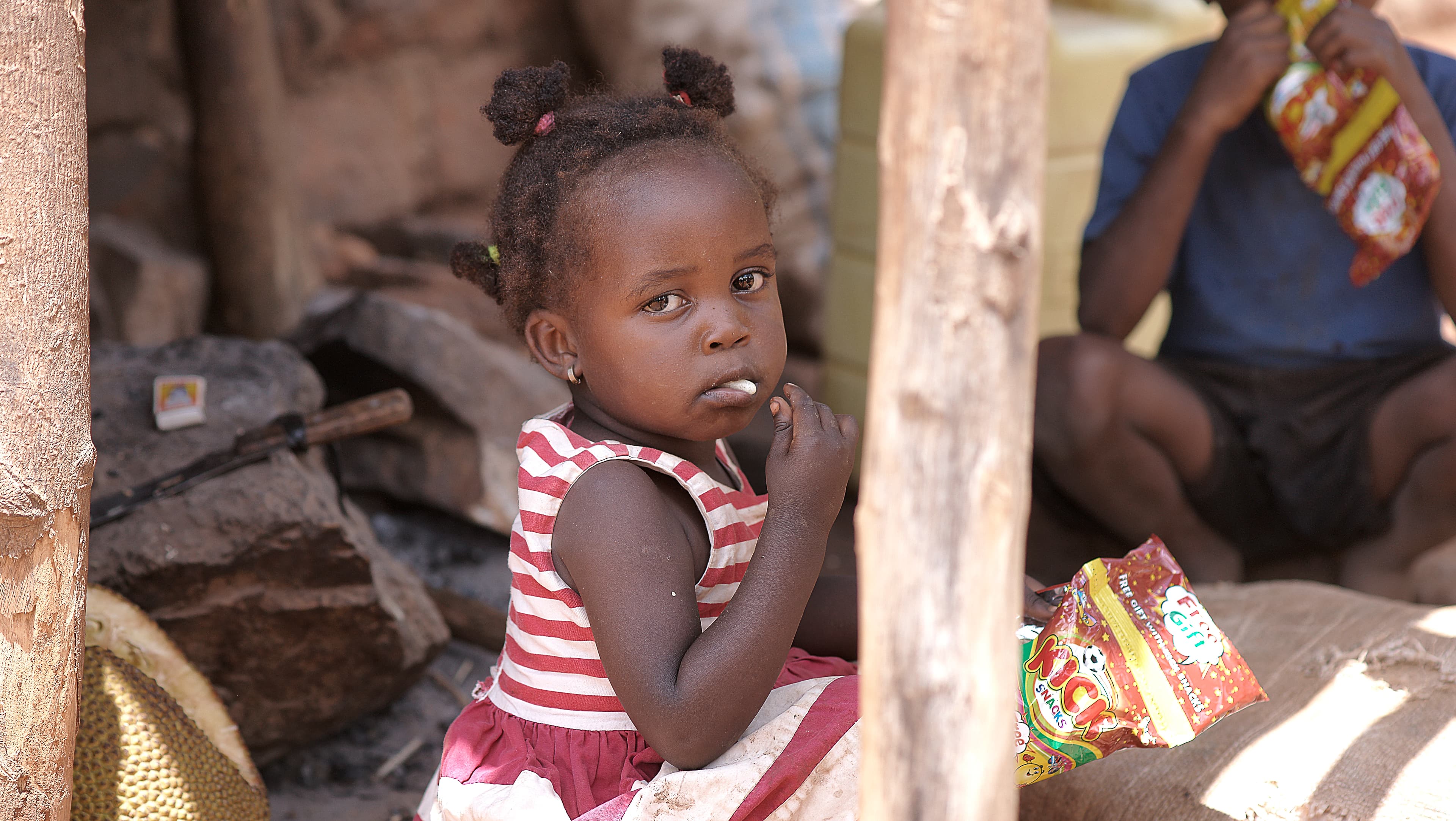 Child in a red-striped dress standing near wooden beams holding a food packet